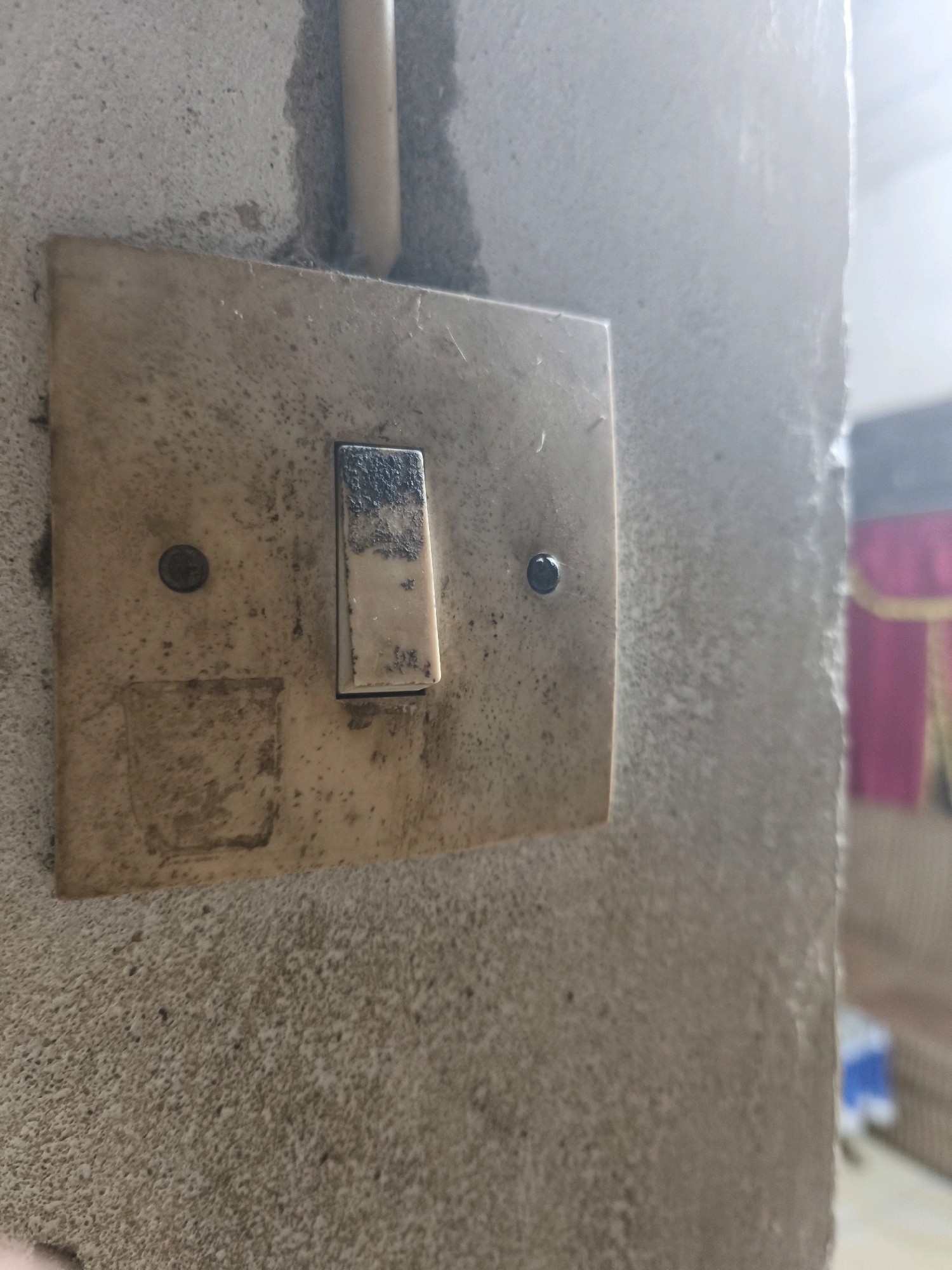 A light switch is mounted on a concrete wall in an interior setting. The switch is a simple toggle switch with a rectangular, off-white plastic cover. The cover is stained and appears to be aged. There is a small, circular screw visible on the left side of the cover. A thin, beige conduit runs vertically behind the switch. The wall itself is textured and appears to be made of rough concrete. To the right of the switch, the wall recedes, and a blurry background shows hints of fabric, possibly curtains or drapes, in shades of pink and gold. The lighting in the image suggests a natural light source from the right, casting a shadow on the left side of the switch. The overall impression is one of an older, functional electrical fixture in a utilitarian environment. The location is identified as Unknown, Indonesia.