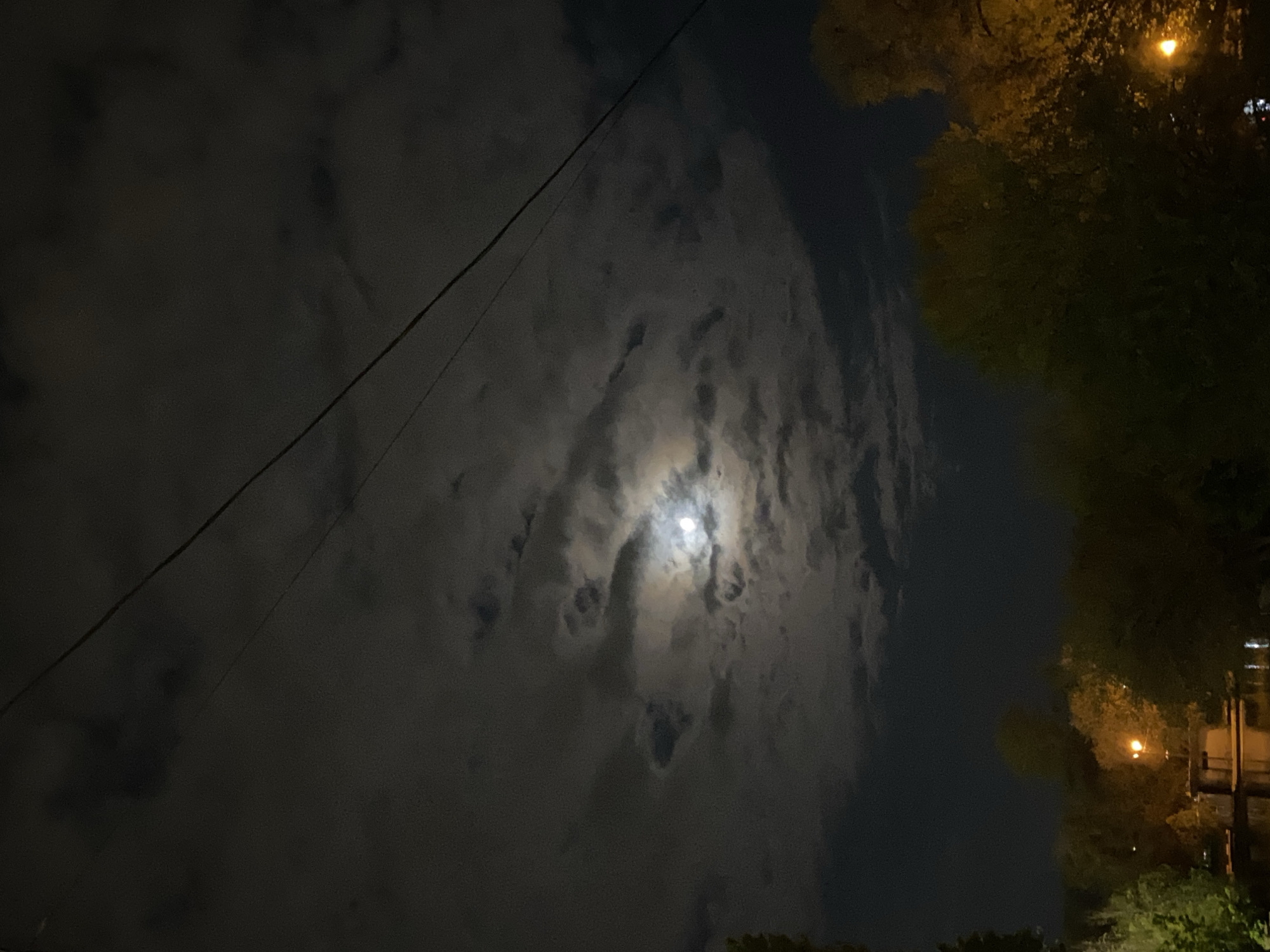 The image captures a nighttime scene, oriented vertically as if looking upwards, showcasing a dramatic interplay between clouds and celestial light. The dominant subject is the sky, filled with dense, dark clouds that cover most of the frame, particularly on the left and central portions. Through a break in these clouds, a bright, full or near-full moon is visible, casting a soft, ethereal glow that illuminates the edges of the surrounding cloud formations.
A thin, dark power line or utility wire diagonally traverses the upper left quarter of the image, extending towards the center. On the right side, the dark silhouettes of trees and foliage are visible, illuminated in parts by artificial light sources. A prominent orange-yellow glow, likely from a streetlight, is present in the top right corner, brightening some of the tree canopy. Further down on the right, additional hints of artificial light and possibly parts of structures or buildings are faintly discernible against the dark background.
The time of day is clearly night. The weather appears cloudy, yet with enough breaks for the moon to be a central focus. There is no discernible activity or event taking place, as it is a static landscape shot. The presence of power lines and streetlights suggests an urban or suburban environment. No text is visible in the image. The overall impression is one of nocturnal quietude, with the moon peeking through a cloudy sky.