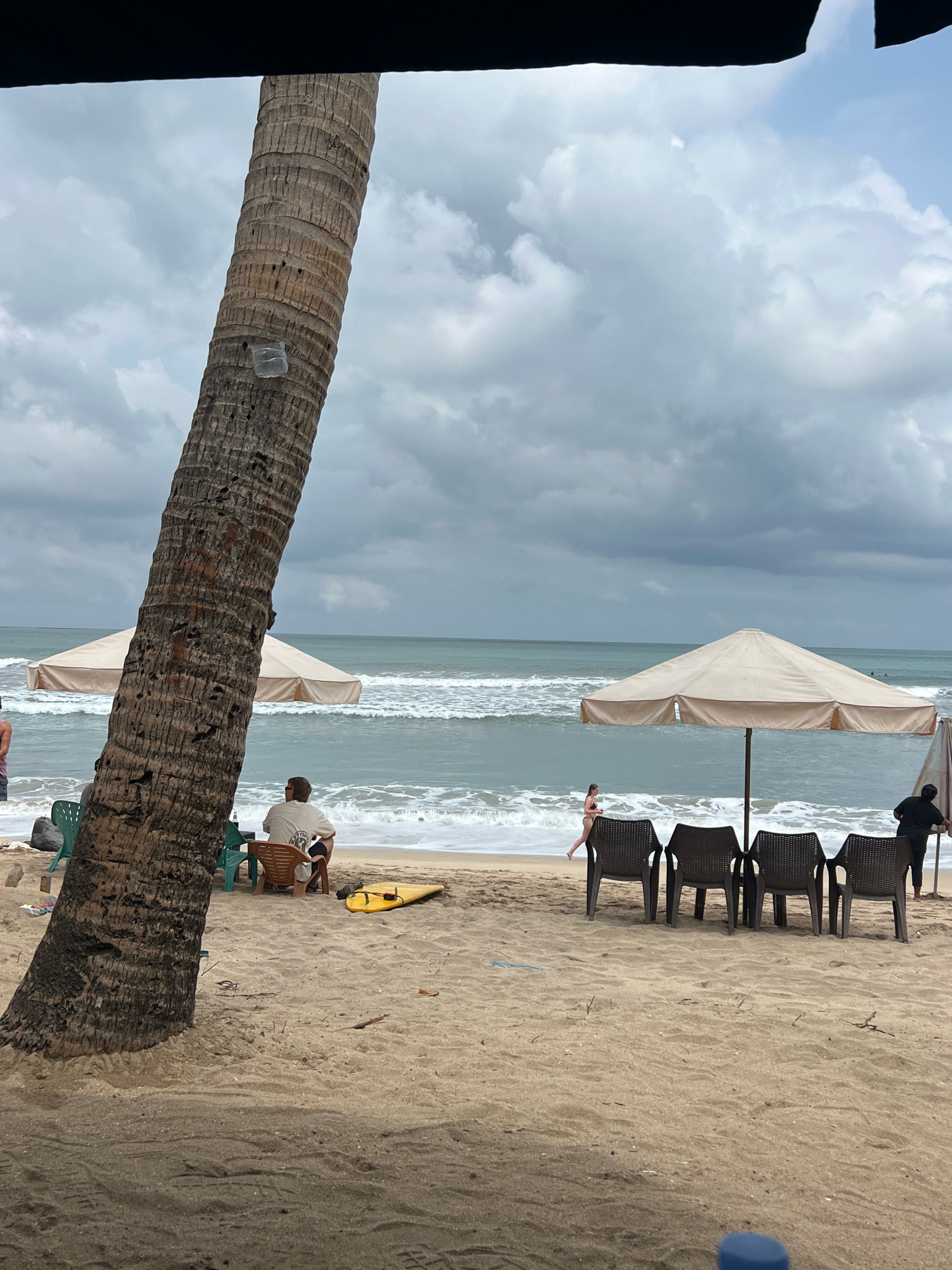 The image depicts a beach scene in Kuta, Indonesia, on an overcast day. In the foreground, a large, textured palm tree trunk dominates the left side of the frame. The beach is sandy, with subtle patterns of sand rippling from the tide.
Several objects are visible on the sand. To the right of the palm tree, a man is seated in a chair, facing the ocean. A yellow surfboard rests on the sand near him. Further to the right, a large, light-colored beach umbrella provides shade over a row of four dark chairs. A young woman in a swimsuit is walking along the edge of the water in the background. Behind her, another person, wearing dark clothing, stands near a smaller umbrella.
The ocean is visible in the midground, with moderate waves breaking on the shore. The sky is filled with heavy, grey clouds, suggesting potential for rain. The overall atmosphere is calm, with a sense of tropical relaxation.