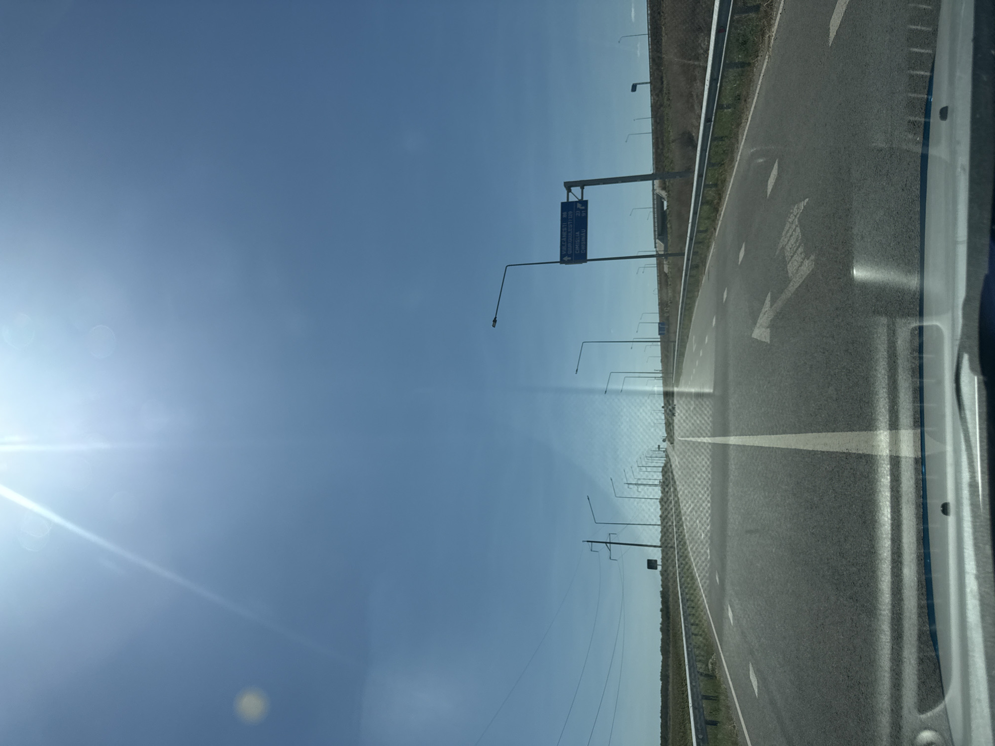 The image shows a view from the perspective of a car driving on a straight, asphalt road under a bright, sunny sky. The road has a white line marking the center and is bordered by a metal guardrail on the right and what appears to be a field of dry grass or crops on the left. There are several streetlights and power lines visible along the sides of the road. In the distance, a blue road sign with white lettering indicates directions to "VULCANESTI 48", "GIURGIULESTI 129", "LEOVO 31", and "CHISINAU 81". The overall environment suggests a rural or semi-rural highway setting. There are no people or animals visible in the image. The sun is high in the sky, causing some glare and lens flare in the upper portion of the image. The image is dated from the perspective of being taken from a vehicle.