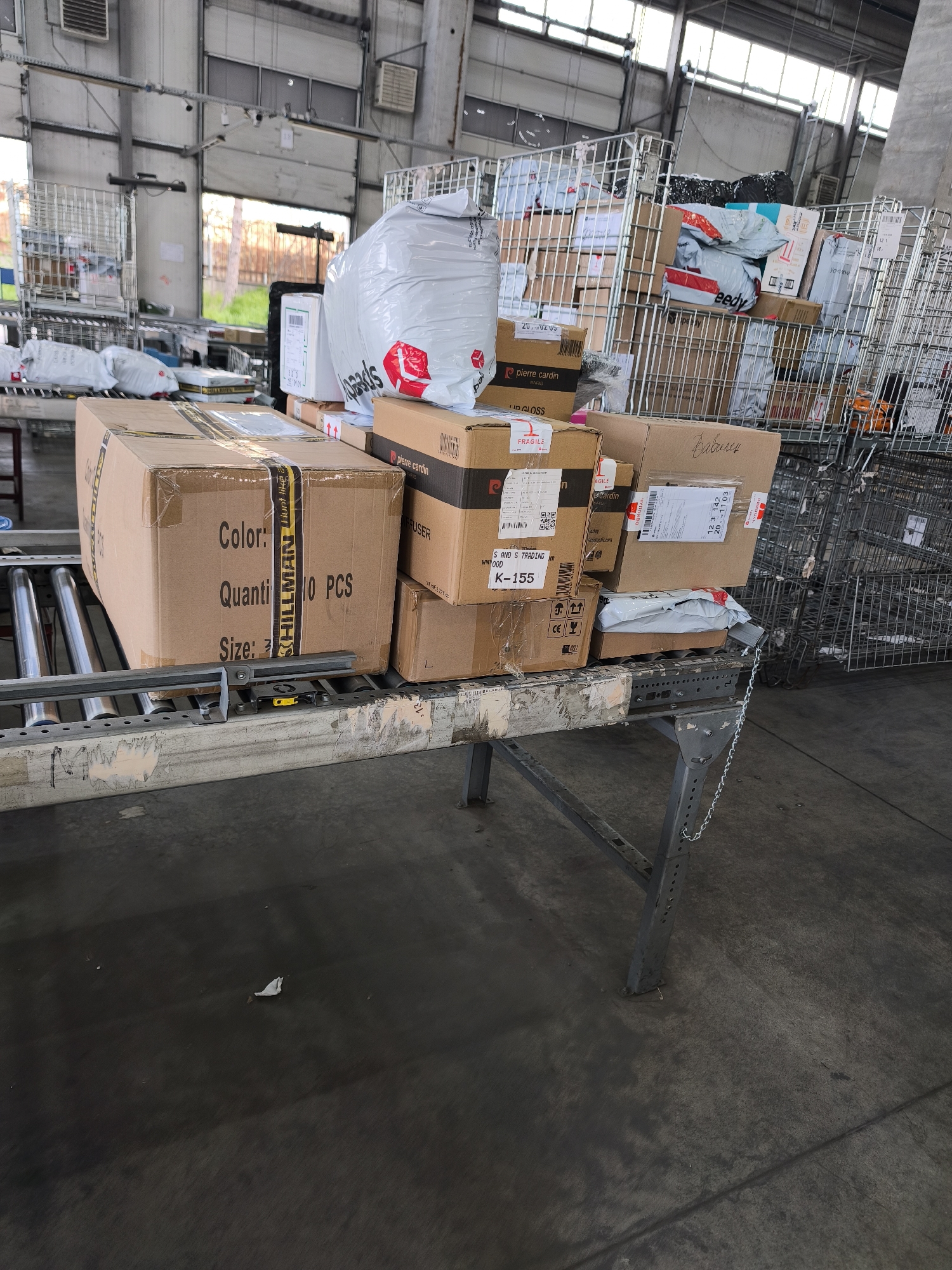 The image depicts an indoor scene within a logistics facility or warehouse, likely a sorting area. No people are visible. The primary subjects are numerous packages, parcels, and cardboard boxes of various sizes, stacked and arranged on a roller conveyor belt system and within metal rolling cages.
In the foreground, a large brown cardboard box rests on the conveyor belt, taped with yellow and black striped tape. To its right, several other boxes are stacked, some bearing the "pierre cardin" brand. A prominent white plastic bag with red branding, possibly from "Speedy," sits on top of these boxes. More packages and boxes, some wrapped in white plastic, are visible further down the conveyor and within the metal cages in the background. The cages are filled with an assortment of parcels, suggesting a high volume of goods in transit.
The setting appears to be a large industrial building with high ceilings and substantial concrete walls or panels. Large windows or garage doors in the background allow natural light to enter, indicating it is daytime. The floor is concrete. The overall impression is one of organized chaos, typical of a busy parcel sorting or distribution center.
Visible text and notable details include:
- On the large foreground box: "Color:", "Quantity 10 PCS," "Size:", and "SHILLMAN Hunt like a" on the yellow tape.
- On several boxes: "pierre cardin," "DIFFUSER," "ID GLOSS," "K-155," "FRAGILE" with an icon, "CE," "AGILE," and "Kosomatic.com."
- On a white bag and small box: "Speedy."
- Handwritten on a box: "Вавилен" (Vavilen).
- On a label with a fragile icon: "УПЛИВО" (Uplivo), likely Bulgarian for fragile.
- Barcode and tracking numbers are visible, such as "12 3 142 20 _ 1103" and "20,0205."
Given the "Speedy" courier branding and Cyrillic text, the scene aligns with the provided location context of Burgas, Bulgaria, indicating a parcel handling operation in the region.