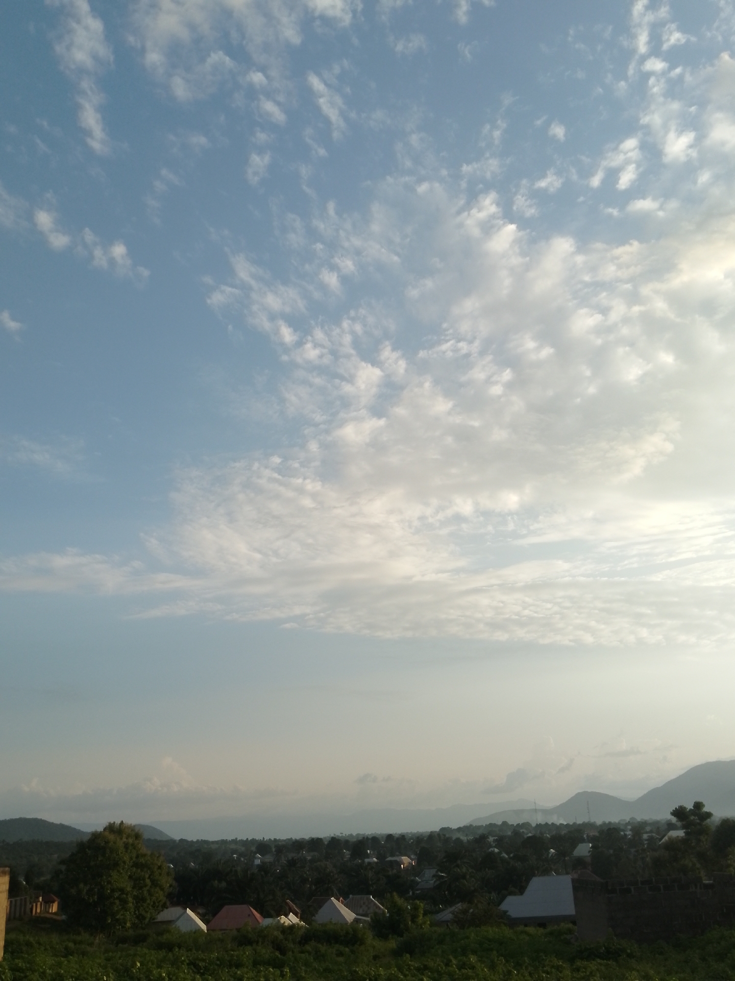 The image captures a panoramic view of a landscape from an elevated position, likely at dusk or dawn. The sky dominates the upper two-thirds of the frame, filled with a scattering of thin, wispy clouds against a pale blue backdrop. The lower third of the image reveals a sprawling settlement nestled amongst lush greenery and rolling hills. Numerous houses with varied roof colors, predominantly white, red, and grey, are visible. Dense foliage, including trees and bushes, intersperses the settlement. In the distance, the silhouette of mountains can be seen against the hazy horizon. The overall impression is one of a tranquil rural or semi-urban environment under a vast, open sky.