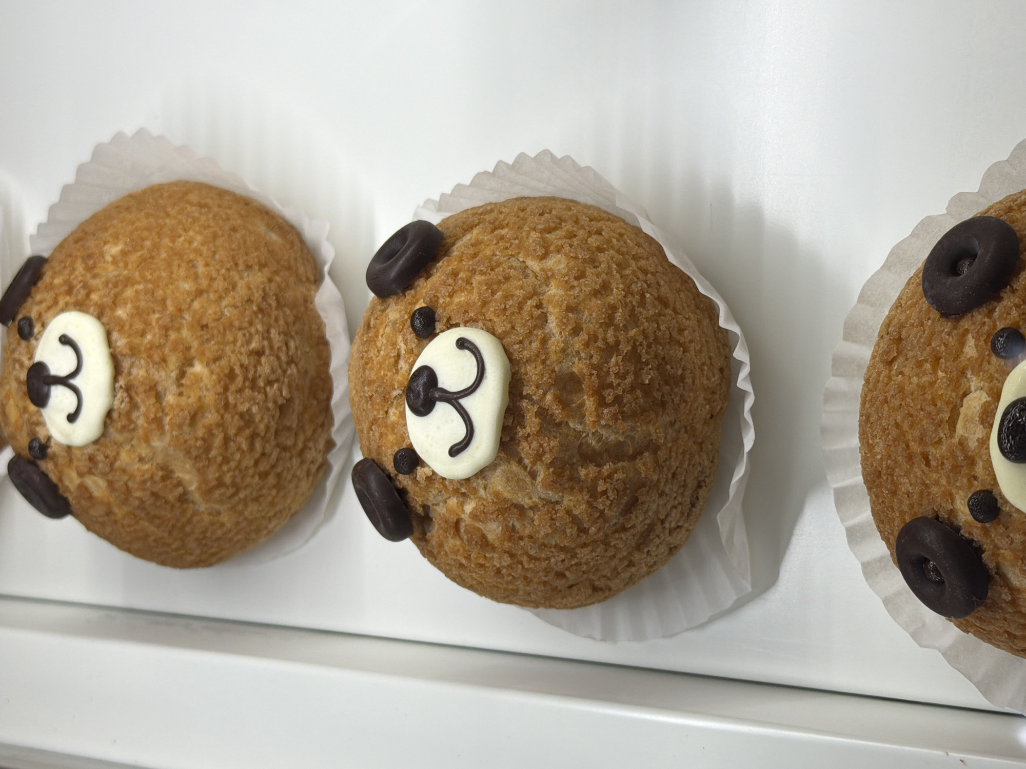 Three cream puffs in white paper liners are arranged in a row on a white surface. Each cream puff is decorated to resemble a bear's face, with dark chocolate pieces used for the ears and eyes, and a white icing snout with a black icing detail for the nose. The scene appears to be indoors, likely a bakery or dessert shop. The precise location in London, United Kingdom, is not discernible from the image.