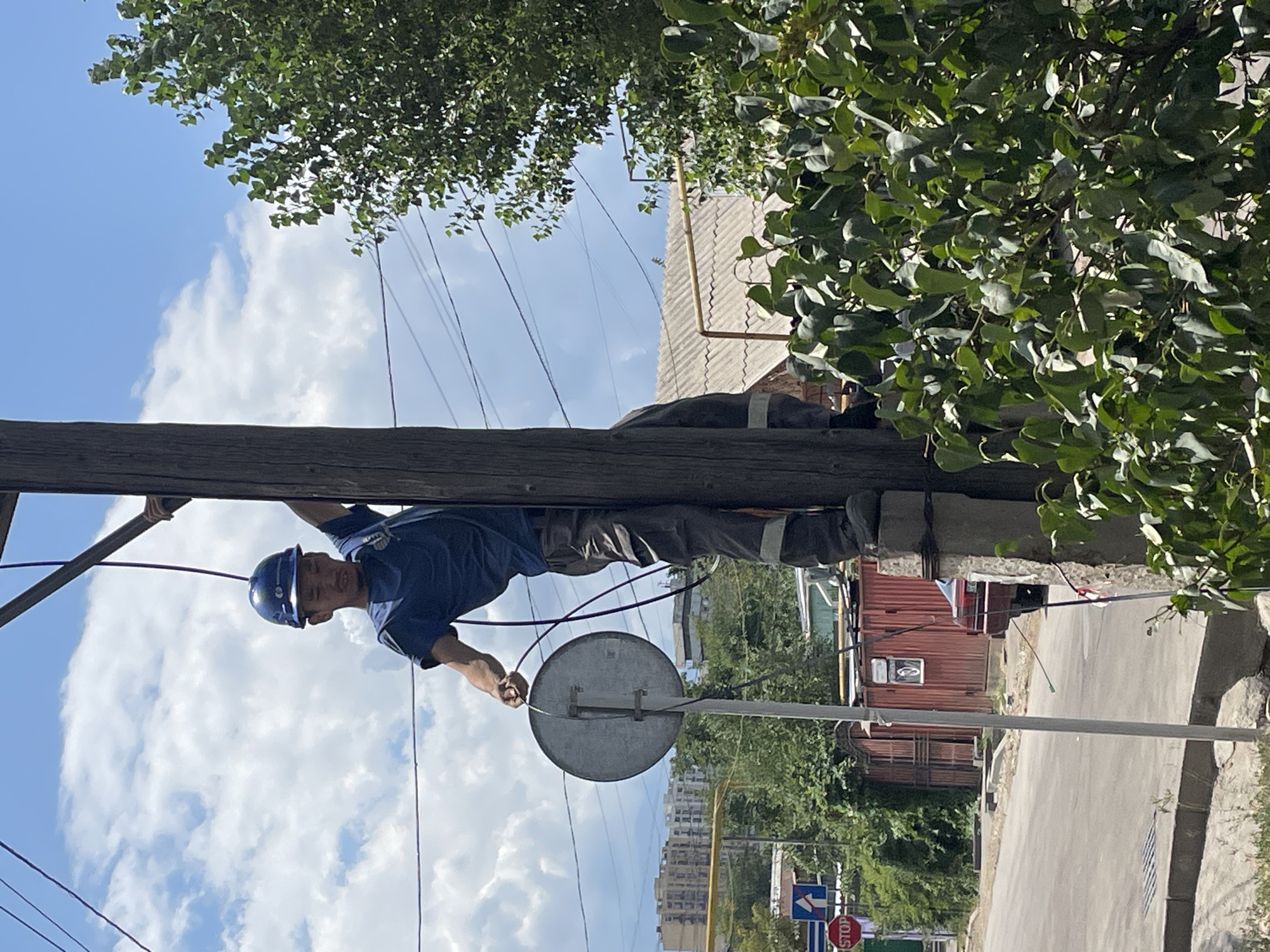 A person wearing a blue hard hat and a blue t-shirt is climbing a wooden utility pole. The person is holding a round metal sign and a dark cable in their hands. The person is wearing gray work pants with reflective stripes on the lower legs. In the background, there are buildings, trees, power lines, and a blue sky with white clouds. To the left of the pole, there is a stop sign and a directional sign. The scene appears to be in a city. The architecture and the presence of specific street signs, such as the stop sign and directional arrow, indicate the location is likely Almaty, Kazakhstan. The overall environment suggests a typical urban street scene on a bright, sunny day.