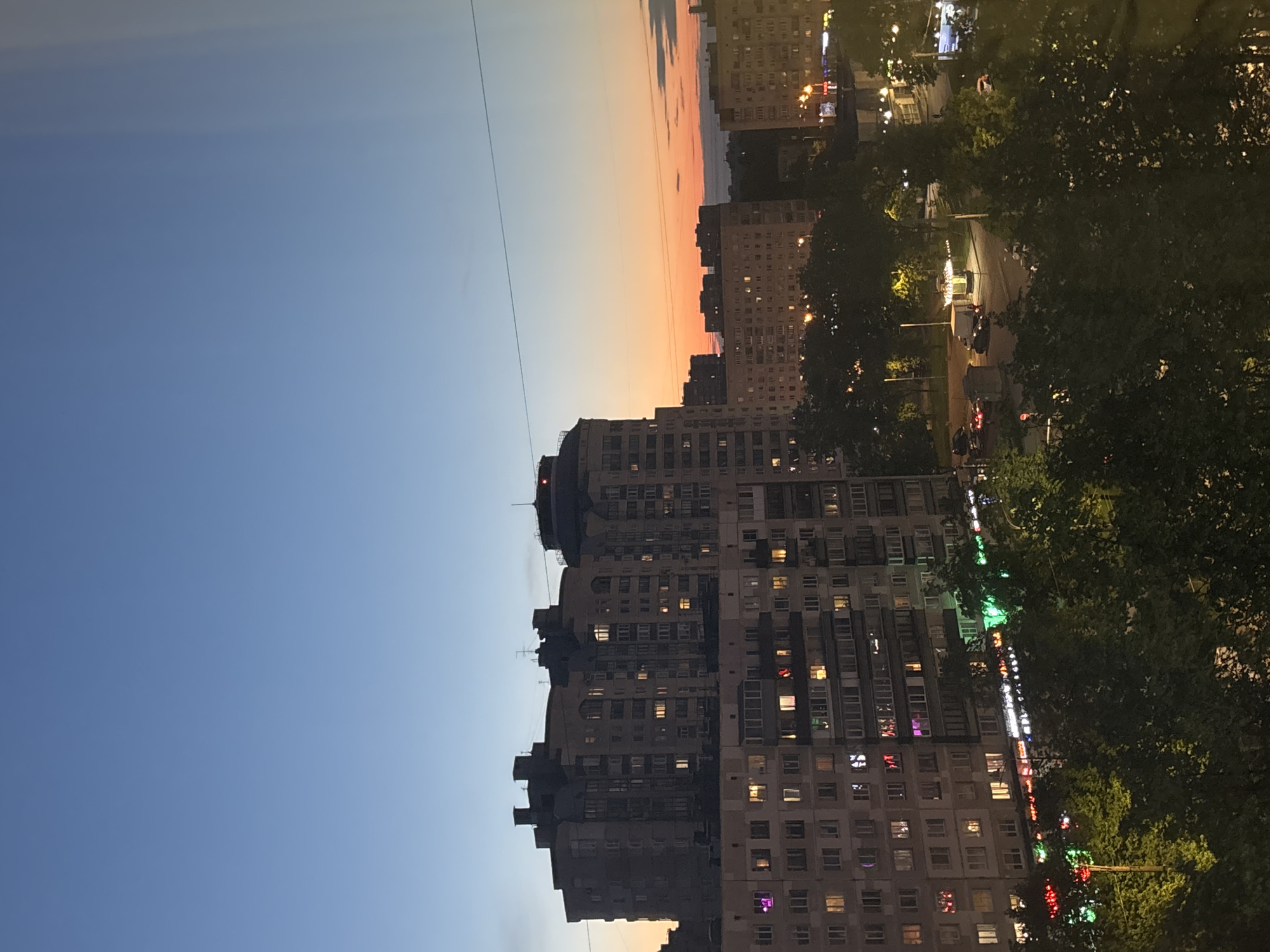 The image depicts a cityscape at dusk, likely in a residential area of Vsevolozhsk, Russia. Several large apartment buildings dominate the scene, with many illuminated windows suggesting residential occupancy. A street with parked cars and occasional moving vehicles is visible, bordered by trees. The sky transitions from a blue hue at the top to a warm orange and pink near the horizon. A single power line stretches across the sky.