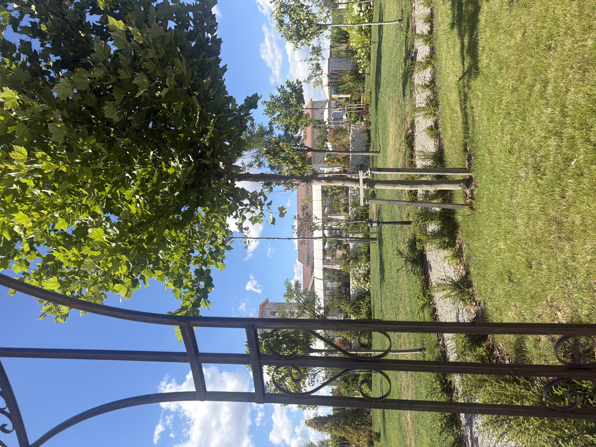 A wide-angle, eye-level outdoor shot captures a lush green lawn in front of a building complex. The foreground is partially obscured by a dark, ornate metal archway. The lawn is well-maintained with some trees planted at intervals. A paved pathway winds through the grass. In the background, a long, white-canopied structure with tables and chairs suggests an outdoor dining area. Behind this, a building with a reddish-tiled roof and a prominent tower is visible. The sky above is a clear blue with scattered white clouds. The scene is peaceful and appears to be a well-landscaped garden or outdoor seating area for a restaurant or event venue. This location is identified as the area surrounding the Konyatska Winery in Kocherinovo, Bulgaria.