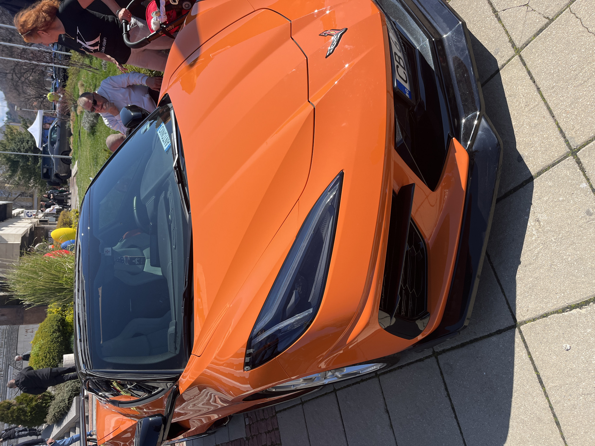 The image shows a close-up of an orange sports car, specifically a Chevrolet Corvette C8, parked on a paved surface. The car is angled in the frame, focusing on the front of the vehicle, including a headlight, front bumper, and hood. A small flag, possibly from Bulgaria, is visible near the front. Several people are visible in the background, including individuals looking at the car. There are also trees, bushes, and buildings in the background. The location appears to be outdoors, possibly a public space such as a park or a street with shops.