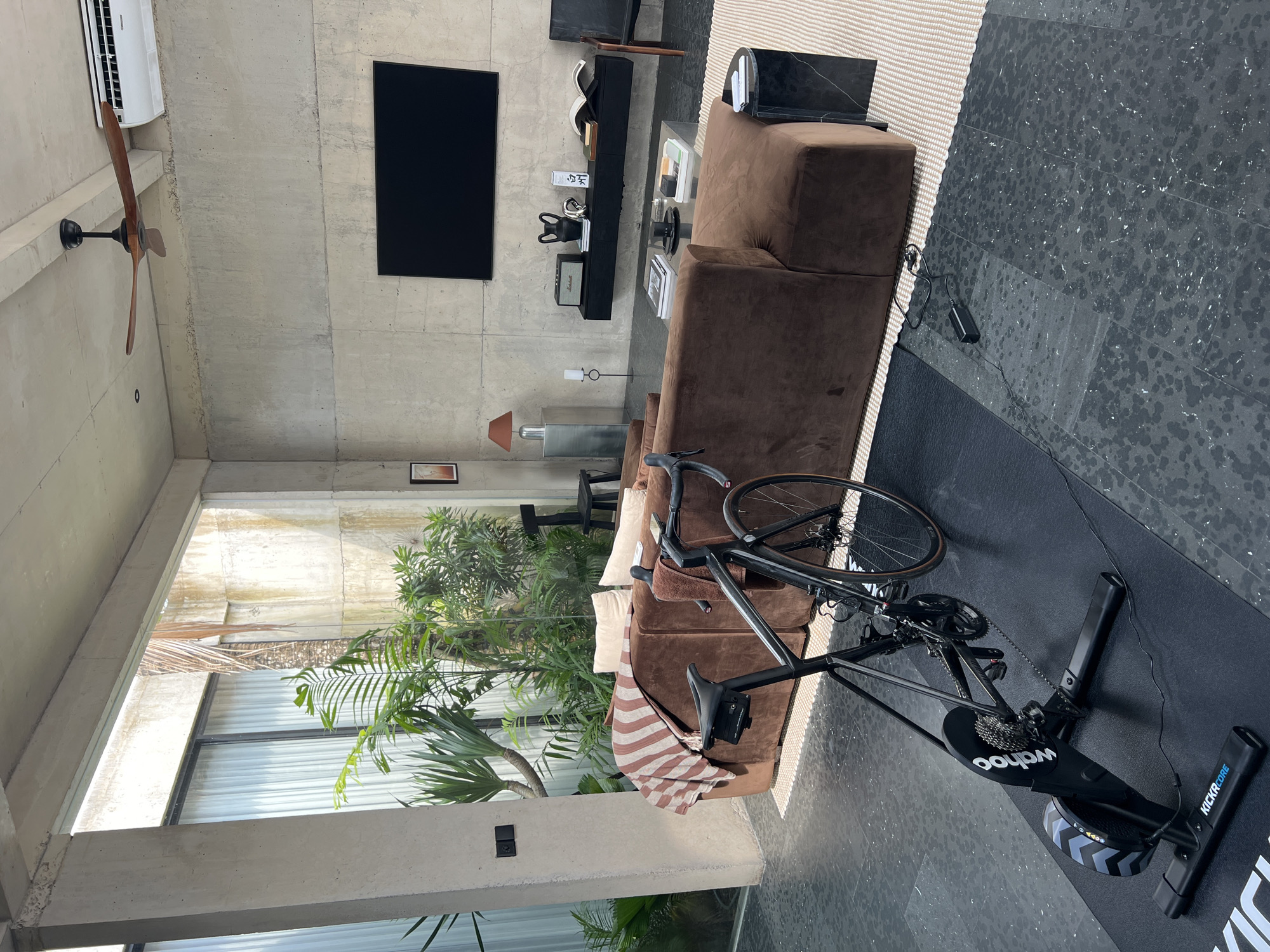 A well-lit, modern living space in Mengwi, Indonesia features a concrete ceiling with a ceiling fan and an air conditioning unit. A large, black flatscreen TV is mounted on the concrete wall above a long, black shelf holding various decorative items. Below the shelf is a table lamp and a small framed picture.
A long, brown sectional couch occupies a significant portion of the room. A bicycle is positioned on a black mat near the couch, set up with a Wahoo Kickr Core indoor trainer.
Outside, a lush green garden is visible through large windows. The floor is covered with dark, speckled tiles, except for a beige rug that runs along the side of the couch. A black marble side table is placed on the rug near the couch.