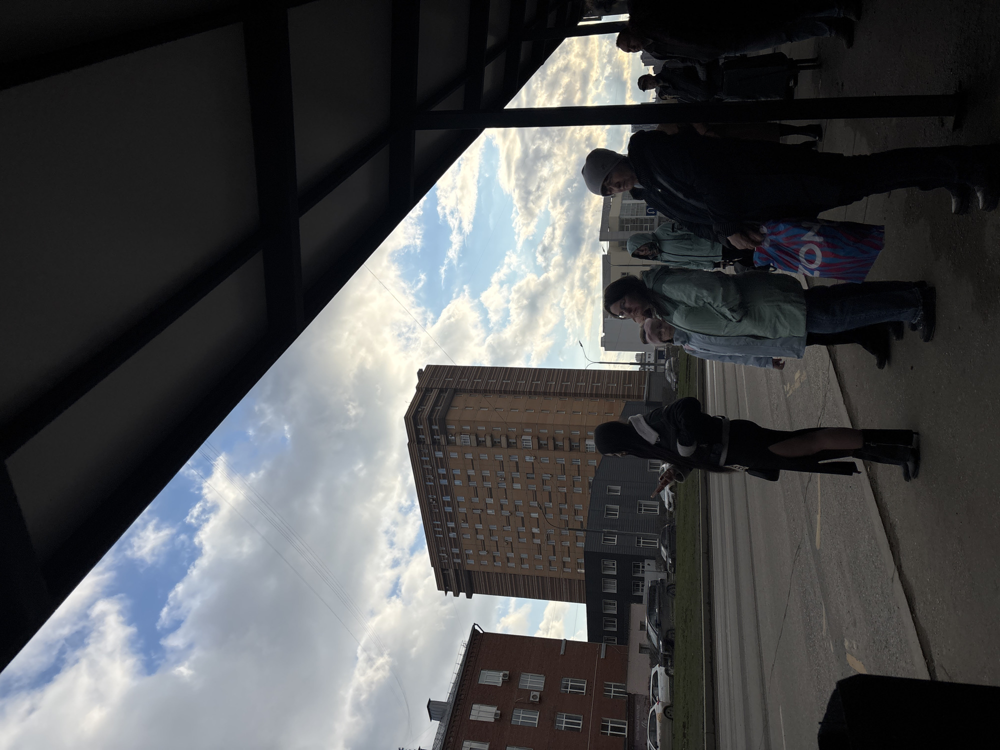 A low-angle view from what appears to be a bus shelter or covered walkway in Perm, Russia. The sky is visible through the opening of the shelter, with clouds scattered across a blue sky. Several people are standing to the right of the frame, facing towards the street. Buildings of varying heights, colors, and architectural styles are visible in the background. Cars are parked alongside the street, and a road is directly in front of the buildings.