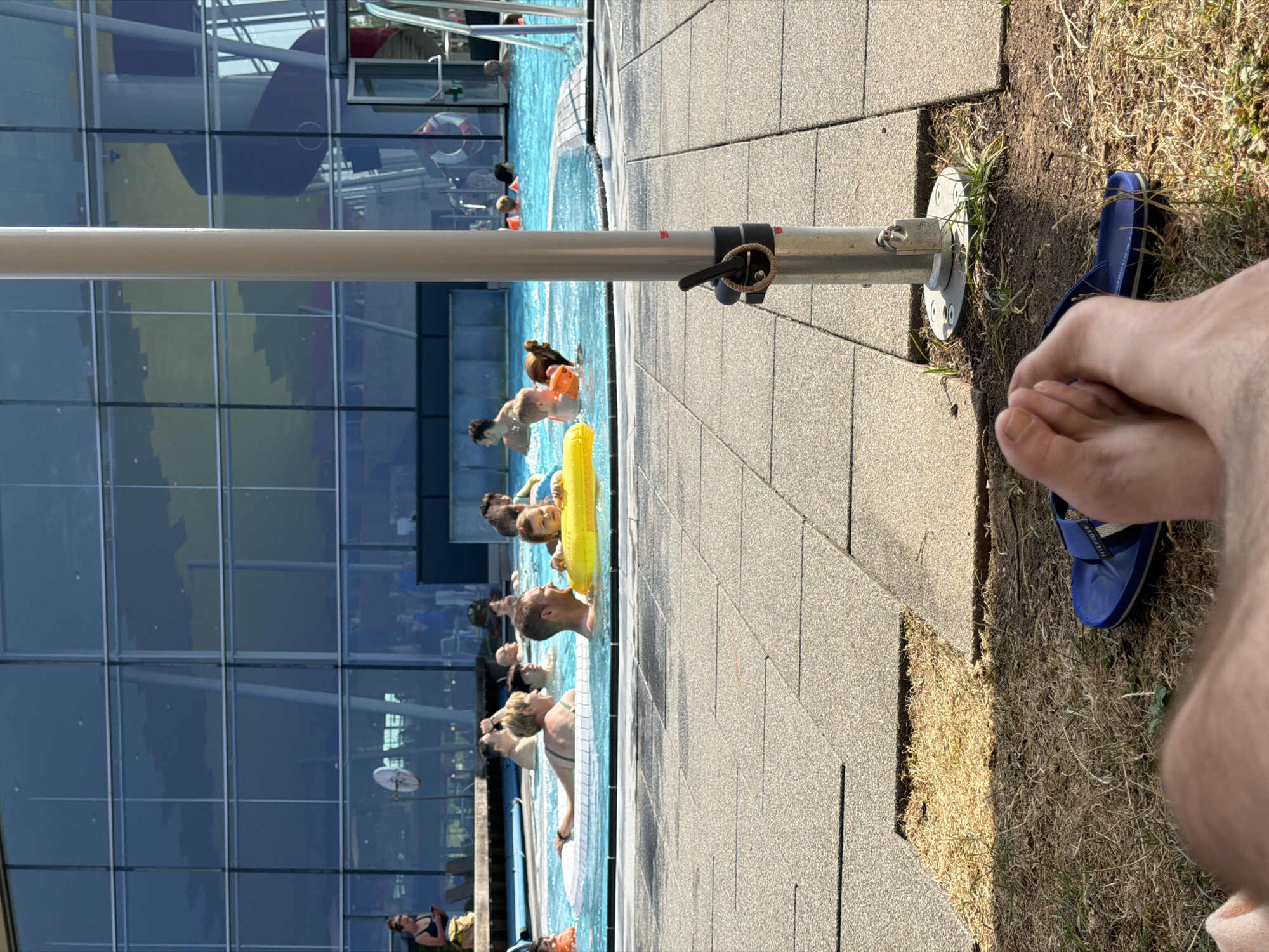 The image depicts a sunny day at a public swimming pool facility, likely the 'Akaplats' aquatic center in Niederanven, Luxembourg, given the architectural style of the building and the general appearance of the facility. In the foreground, a person's bare feet and a blue flip-flop are visible, resting on dry grass and paving stones. The background features a large, modern building with a glass facade reflecting the sky and surrounding environment.
The main focus of the image is the swimming pool area where numerous people are engaged in various water activities. Several individuals are seen in the clear blue water, some swimming, others relaxing on inflatable rings. Children are present, with one wearing an orange flotation device and another holding a yellow ring. Adults are also visible, some interacting with children, others conversing. The pool area is surrounded by grey, textured paving stones. A prominent silver pole, likely part of a shade structure, stands in the middle ground, with a black clamp attached to it. In the distance, a red and purple water slide is visible within the building, indicating it's part of a larger aquatic complex. The overall atmosphere appears to be one of leisure and recreation on a bright, warm day.