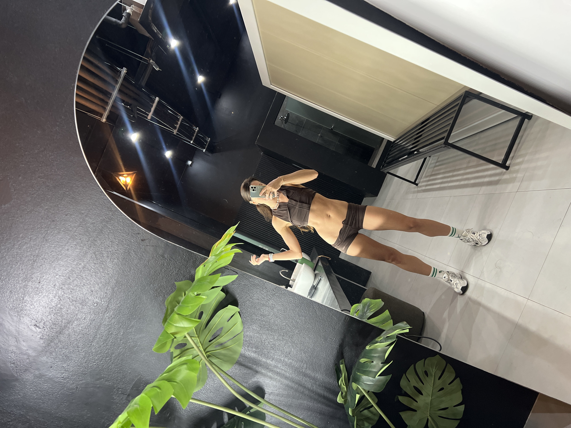 A full-body, mirrored selfie shows a woman in a brown sports bra and shorts standing in a gym locker room. She is holding her phone up to take the picture and has her left arm raised. The locker room has a black textured wall, a large arched mirror, a sink and vanity, a shower stall, and a black metal bench. There is a large artificial plant with green leaves in the foreground. The floor is tiled in white. The ceiling is black and has exposed pipes and lighting fixtures.