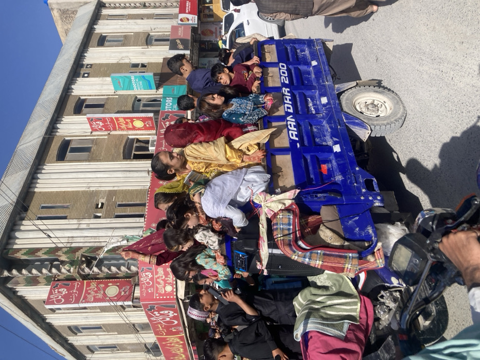 This image depicts an urban street scene in Quetta City Tehsil, Pakistan, observed under clear daylight conditions. The primary subject is a blue three-wheeled motorized cargo rickshaw, branded "AANDAR 200," positioned on an asphalt road. Approximately 15-20 individuals, predominantly children and young adolescents, are seated and standing in the open cargo bed of the vehicle.
In the left foreground, the handlebars and hands of a person operating a motorcycle are visible, indicating the perspective from which the photograph was taken. Draped over the front-left side of the blue vehicle are various fabric items, including a green and white striped blanket and a red and black plaid blanket. A portable speaker or audio device rests on the front interior ledge of the cargo bed.
Among the occupants of the blue vehicle:
* A young boy in a black shalwar kameez and a white cap stands on the far left, holding a small black object resembling a microphone or speaker.
* Several girls are dressed in colorful attire, including yellow, maroon, and light blue traditional garments. Some display henna patterns on their hands.
* One girl in a maroon outfit is visible raising her right arm above her head.
* The individuals in the vehicle appear engaged, with varying expressions suggesting observation, interaction, or participation in an event.
In the background, a multi-story building with a light-colored, vertically ribbed or striped facade is prominent. Commercial signs are affixed to this building and adjacent structures:
* Multiple red banners with white script, translating to "Qabayel Restaurant," are visible, some featuring a plate and cutlery icon.
* A blue and white sign reads "DOCTORS MEDICAL STORE" with a red cross emblem.
* Below it, a smaller black sign displays "WORLDS MOBILE."
* Other indistinct commercial signage is also present.
On the far right, a partial view of a white sedan is visible behind the blue vehicle, and a male pedestrian in a brown shalwar kameez is walking on the roadside. Overhead utility wires traverse the clear blue sky. Shadows cast on the road indicate direct sunlight.