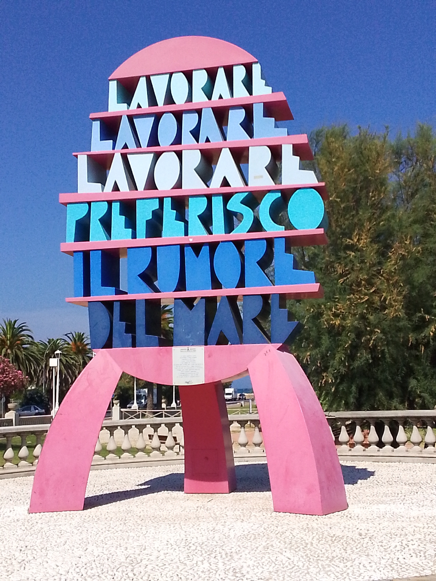 This media file features a vibrant, multi-layered sculpture on a sunny day. The sculpture is primarily pink, with stacked segments of text rendered in light blue and white. The text reads "LAVORARE", "LAVORARE", "LAVORARE", "PREFERISCO", "IL RUMORE", and "DEL MARE", which translates from Italian to "to work", "to work", "to work", "I prefer", "the noise", and "of the sea". This text is arranged in a tiered, architectural style, topped by a pink semi-circular element. The sculpture is supported by two large, curved pink legs that rest on a surface covered in small white pebbles.
In the background, lush green trees and palm trees are visible, suggesting a warm climate. Beyond the trees and sculpture, a light-colored railing with decorative elements runs along the edge of a paved area. A few vehicles and people are present in the middle ground and background, indicating this is likely a public space. The sky is a clear, bright blue, suggesting a midday or afternoon time. A small plaque is affixed to the central support of the sculpture, but the text on it is not clearly legible. The overall impression is one of a colorful and inviting outdoor art installation in a coastal Italian town, possibly San Benedetto del Tronto, as suggested by the context.