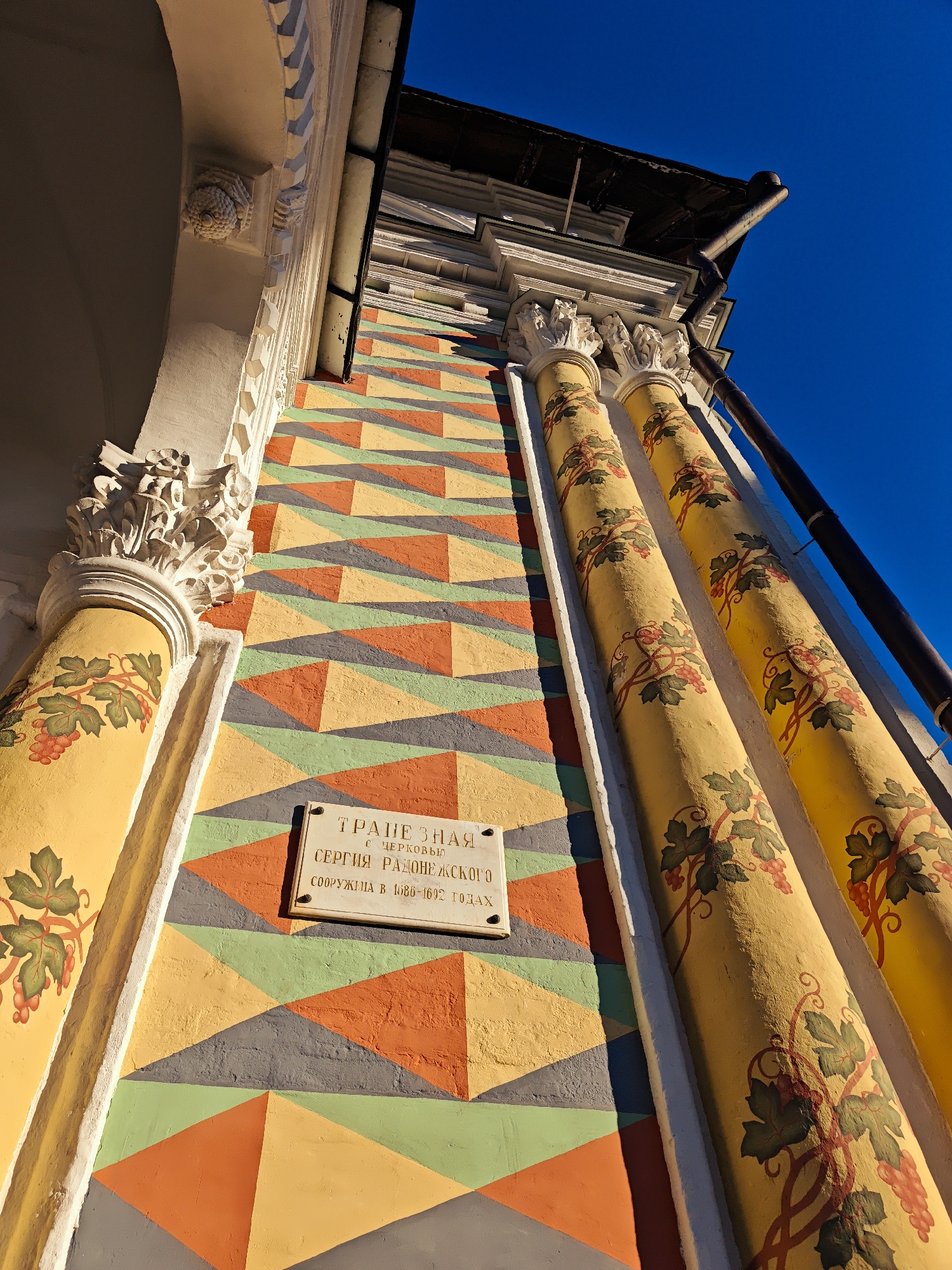 The image is a low-angle shot of an architectural facade against a bright blue sky. The facade features a section with a colorful geometric pattern of triangles in shades of orange, yellow, green, and grey. To the right of this patterned section are two fluted columns painted yellow and decorated with painted green grapevines and clusters of grapes.
Attached to the patterned wall is a rectangular plaque with visible text. The text appears to be in Cyrillic script. It reads: "ТРАПЕЗНАЯ С ЦЕРКОВЬЮ СЕРГИЯ РАДОНЕЖСКОГО СООРУЖЕНА В 1686-1692 ГОДАХ". This translates to "Refectory with the Church of Sergius of Radonezh Built in 1686-1692".
On the left side of the image, a portion of an arched structure with decorative carvings is visible. The sunlight suggests it is daytime, likely morning or afternoon, given the distinct shadows and the intensity of the blue sky, indicating clear weather. The overall impression is one of historical architecture, possibly a religious building given the inscription.