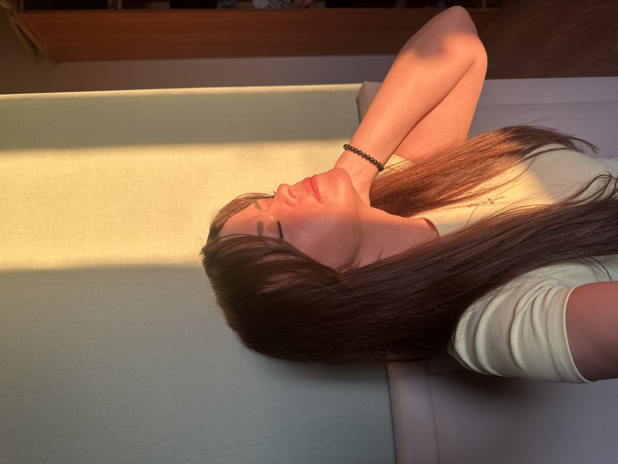An intimate, high-angle shot captures a young woman lying relaxed on a light green or beige upholstered surface. Her long, dark brown hair spreads out behind her. She is wearing a pale yellow or cream long-sleeved top and has a black beaded bracelet on her right wrist. A faint golden cross necklace is visible on her chest. Her arms are raised, with hands clasped behind her head, indicating a state of rest or contemplation. Her eyes appear closed or cast downwards, and her expression seems serene.
Warm, golden sunlight streams into the room, falling directly on her face and the surface, creating bright highlights and a soft, ethereal glow, suggesting it is either early morning or late afternoon. This directional light creates a distinct pattern of illumination across the fabric she is resting on. In the background, out of focus, a wooden shelf or piece of furniture is visible. The indoor setting appears to be a home, possibly a bedroom or living room. The overall mood is one of peace and tranquility. There is no visible text in the image. This scene is set in Chaikovsky, Russia.