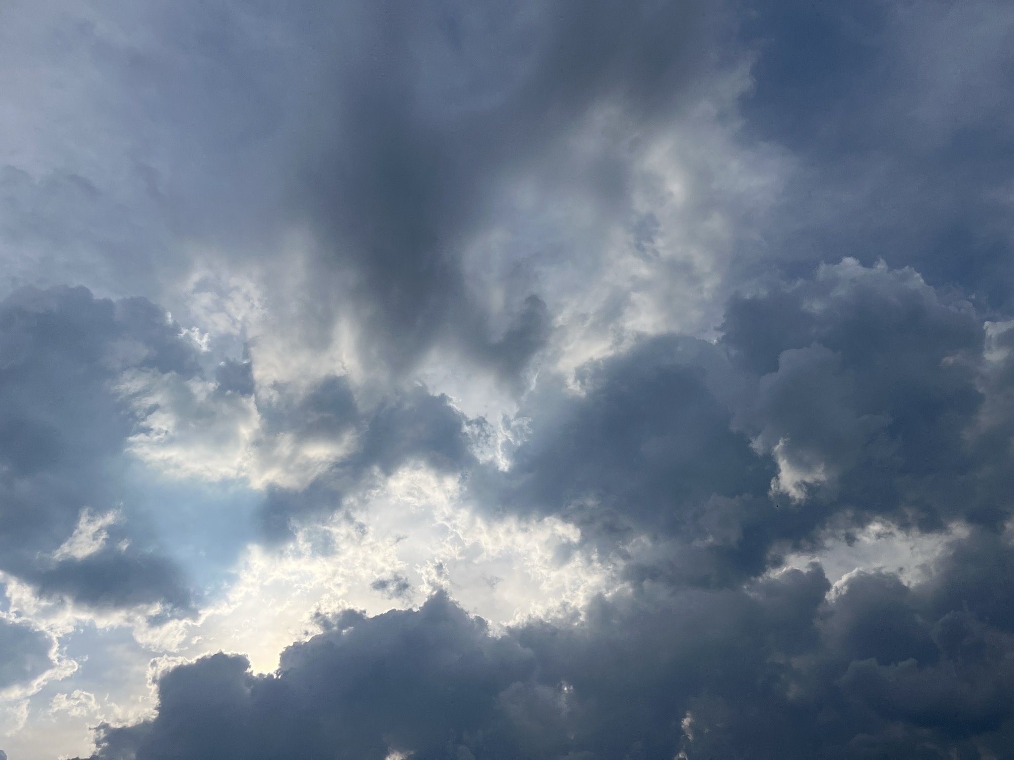 A dramatic, overcast sky with a mix of dark, heavy storm clouds and lighter, illuminated clouds is depicted from a ground-level perspective in Jabłonna, Poland. The sun is mostly obscured, but shafts of light pierce through breaks in the clouds, creating a contrast between the dark and bright areas. The scene is entirely composed of the sky, with no other elements visible. The lighting suggests it could be late afternoon or early evening, or prior to or during a rain shower.