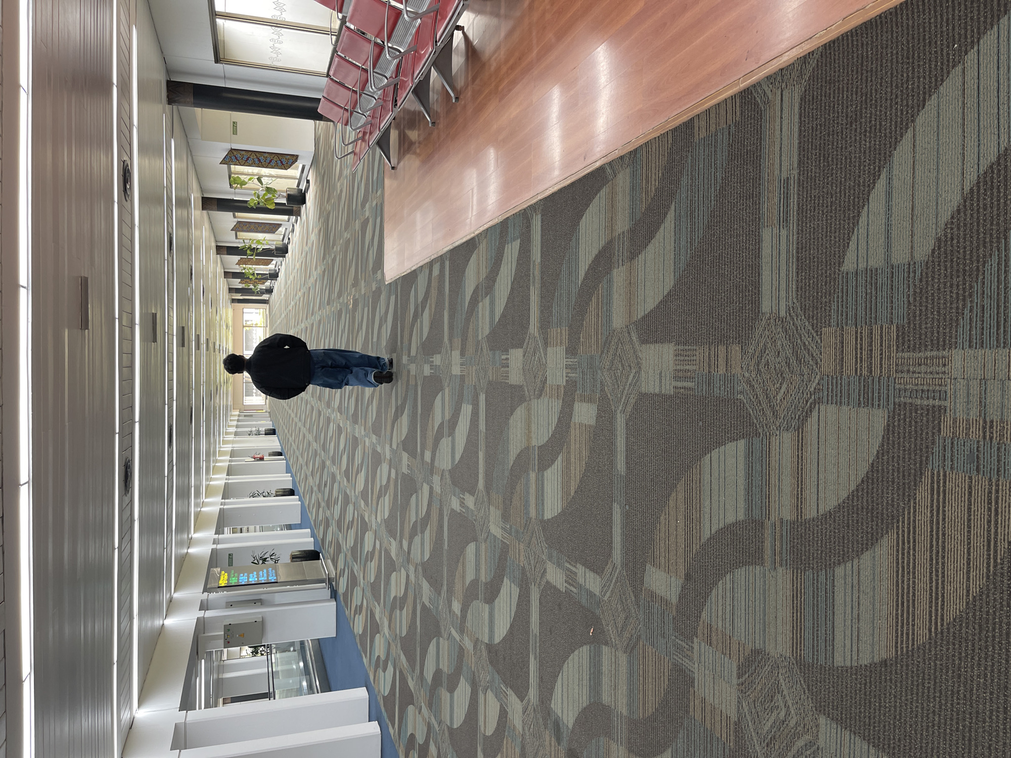 An individual walks down a long, carpeted airport concourse in Tangerang, Indonesia. The concourse features patterned carpet in shades of brown, grey, and blue, with a blue tiled area to the left. Along the right wall, red airport seating is arranged, adjacent to a polished wooden walkway. Fluorescent lights illuminate the space from above, and various signs and potted plants are visible along the walls. The individual, wearing a dark jacket and blue jeans, is walking away from the camera towards the distant end of the concourse.