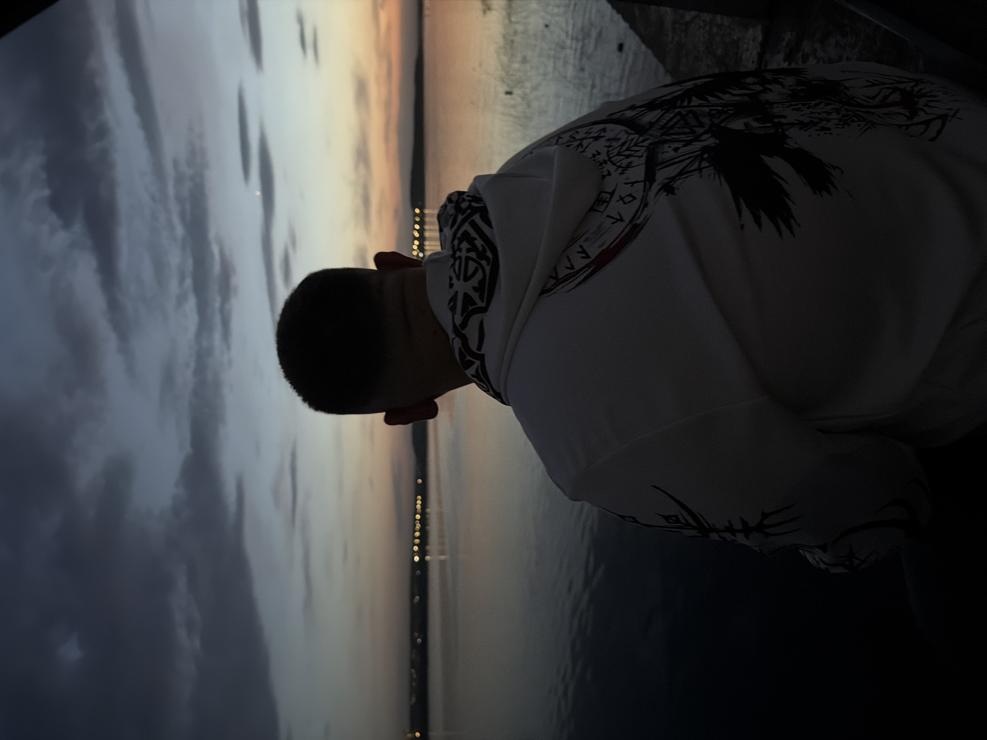 A person with short dark hair is seen from the back, wearing a white hoodie with dark intricate designs, possibly tribal or Norse-inspired, on the hood and sleeves. The person is positioned near a body of water, likely a river or lake. Across the water, lights are visible, indicating a city or town. The sky is divided between dark, cloudy areas and lighter, orange-tinged areas, suggesting either dawn or dusk. Given the prompt's reference to Izhevsk, Russia, it's plausible that the body of water is the Izh River or a reservoir located within or near the city. The person appears to be looking out at the water and the cityscape.