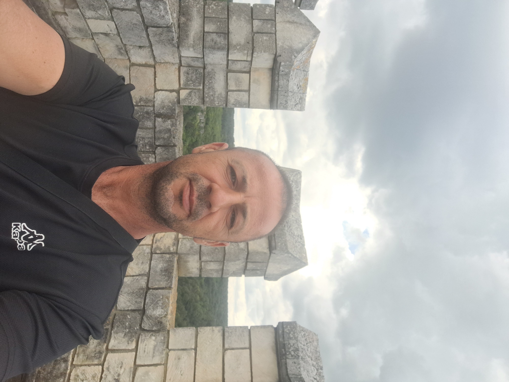 A selfie taken from a slightly high angle shows a middle-aged man with short, dark hair and a stubble beard. He is wearing a black t-shirt with a white Kappa logo on the left side of the chest. The man is standing on what appears to be a stone battlements with crenellations, suggesting a historical site or a fortress. Behind him, a lush green forest stretches into the distance under a cloudy sky. The man is looking directly at the camera with a neutral expression. The setting is likely the Shumen Fortress, a historical monument located in Shumen, Bulgaria, known for its preserved medieval structures and scenic views.