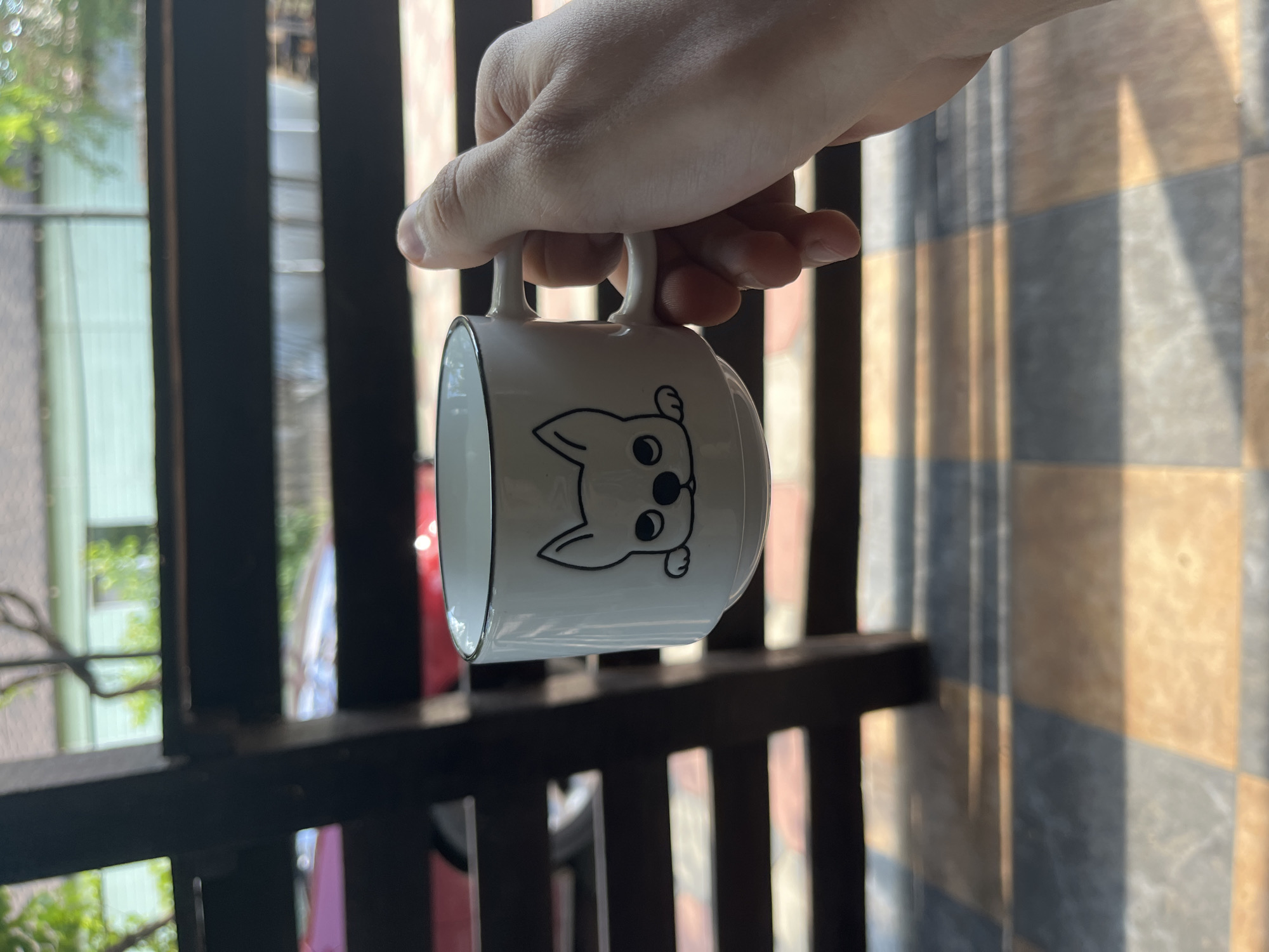 A hand is holding a white ceramic mug with a black outline of a dog's face on it. The mug has a black rim and a white handle. The background is blurred and shows a green building and trees. The scene appears to be outdoors on a patio with a tiled floor. The location is Bohunove, Ukraine.