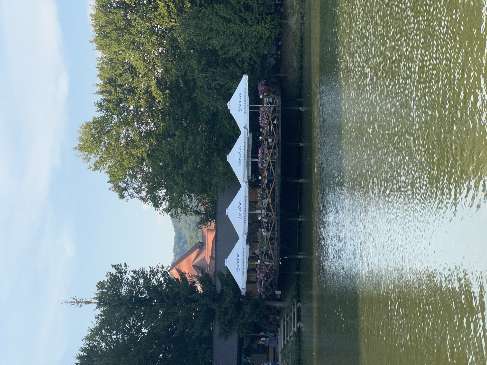 An exterior shot captures a waterside establishment with a deck and umbrellas. The establishment is situated on the edge of a body of water. The deck is furnished with tables and chairs and is adorned with floral arrangements. Four white umbrellas, each bearing the "Gaaarden" logo and the phrase "THE GAARDEN IS OPEN," provide shade over the deck. Several people are visible on the deck, some seated and others standing. A set of wooden stairs leads from the shore to the deck on the left side of the image. A large building with a reddish-brown roof is visible behind the deck, partially obscured by trees. The background is filled with lush green trees and a distant hillside. The sky above is a light blue with scattered clouds. The water in the foreground is calm, reflecting the sky and the surrounding greenery. The scene appears to be set in the city of Сонячне, Ukraine, suggesting a recreational or dining area by a lake or river.