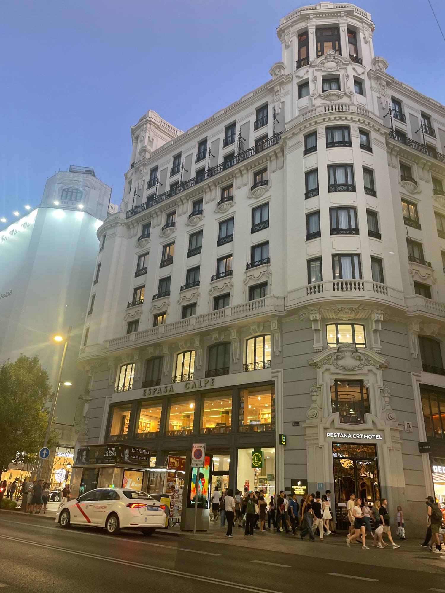 In Madrid, Spain, on a street corner at dusk, a tall, ornate white building with multiple stories dominates the scene. The building features balconies with black railings, decorative architectural elements, and numerous windows, some illuminated from within. The top of the building culminates in a rounded turret-like structure. To the left, a significantly larger, lighter-colored building is partially obscured, its surface appearing to be a large advertisement or screen displaying text.
In the foreground, a white taxi with "FREENOW" branding is parked on the street. A group of people, dressed in casual attire, are walking on the sidewalk in front of the buildings. Some are looking at their phones, while others are engaged in conversation. A street lamp with a warm glow is visible on the left side of the image, casting light on the street and sidewalk. Signs are visible on the buildings, including "ESFASA CALPE" on the lower level of the white building and "PESTANA CR7 HOTEL" above an entrance. The sky is a clear twilight blue.