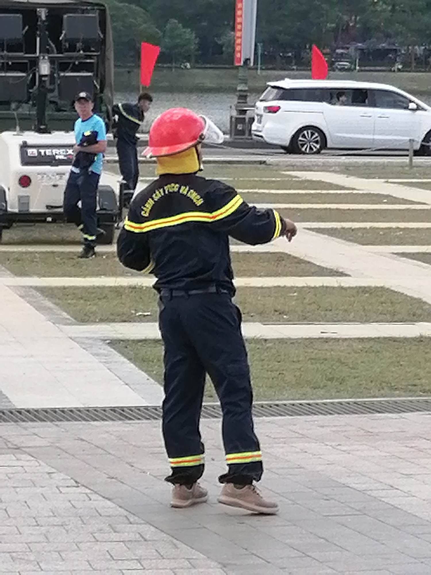 The image shows a street scene in Da Lat, Vietnam. In the foreground, a person wearing a dark uniform with yellow and red stripes on the sleeves and pant legs, and a red helmet, is facing away from the camera, gesturing with their right hand. The uniform has Vietnamese text on the back. In the middle ground, to the left, another person wearing a blue shirt and a black cap is standing near a white trailer with the brand "TEREX" visible. Behind them, a white minivan is parked. Red flags are visible on poles in the background. The scene appears to be outdoors, with paved ground in the foreground and grass in the background.