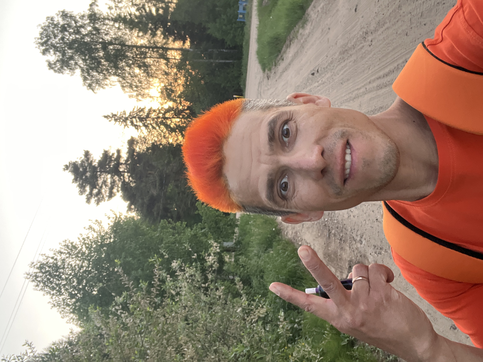 A middle-aged man stands on a dirt road in what appears to be a rural or semi-rural location in Chaikovsky, Russia. He has short, graying hair with the top portion dyed bright orange. He wears an orange t-shirt with black trim. The man is making a peace sign with his left hand, which also holds a small, dark-colored object. He wears a wedding ring on his ring finger.
The environment consists of a dirt road with tire tracks. The surrounding area includes greenery, bushes, and trees. The sky is bright, possibly indicating sunrise or sunset. A blue structure can be seen in the distance.