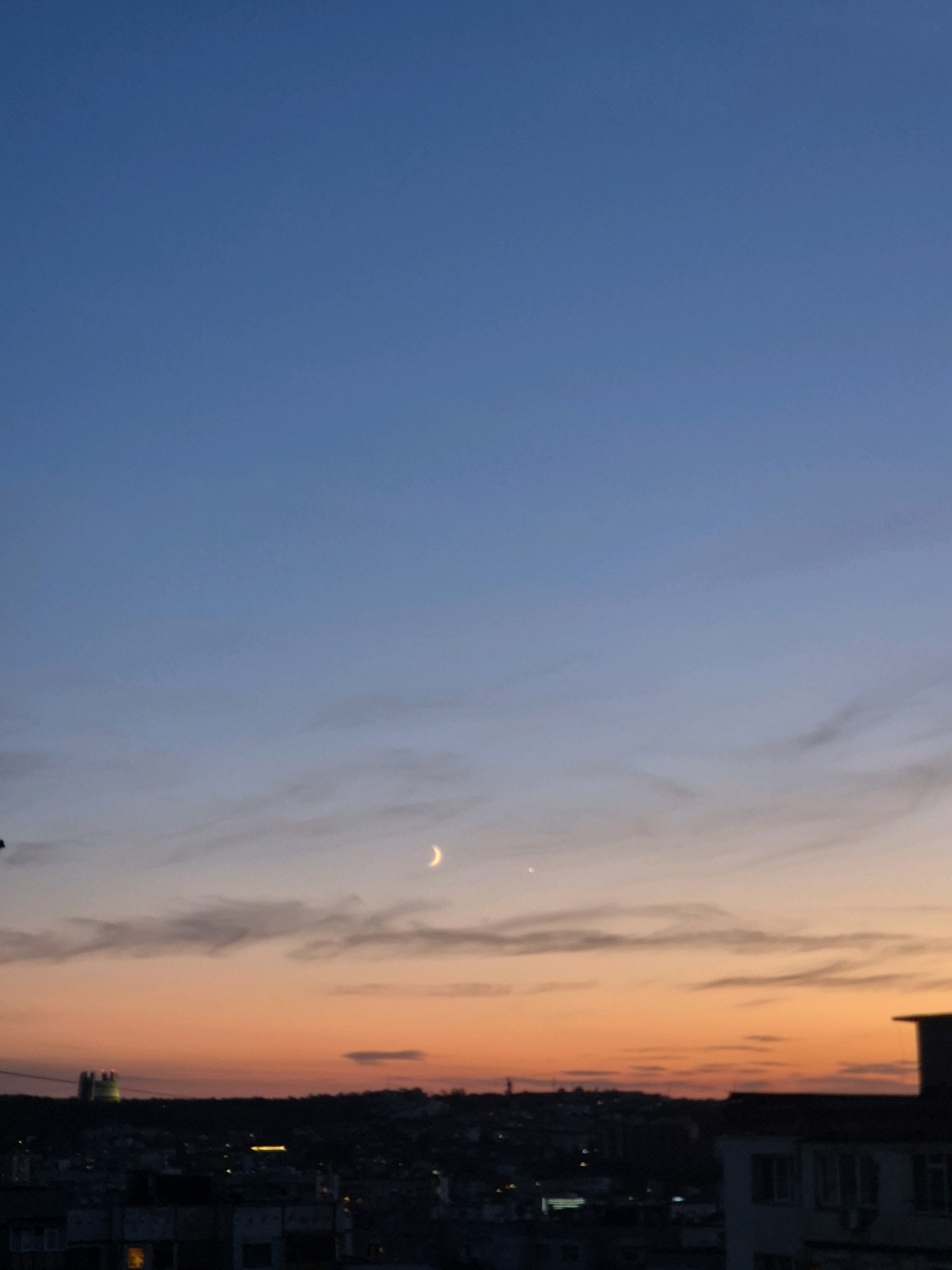 An outdoor, vertical, medium shot captures a city skyline at dusk. The sky transitions from a deep blue at the top to a warm orange near the horizon. A crescent moon and a bright star are visible in the upper portion of the sky. The silhouette of a city with buildings and faint lights forms the bottom half of the image, with a distinct structure resembling a castle or tower on the left side. The lighting suggests it is either sunrise or sunset.