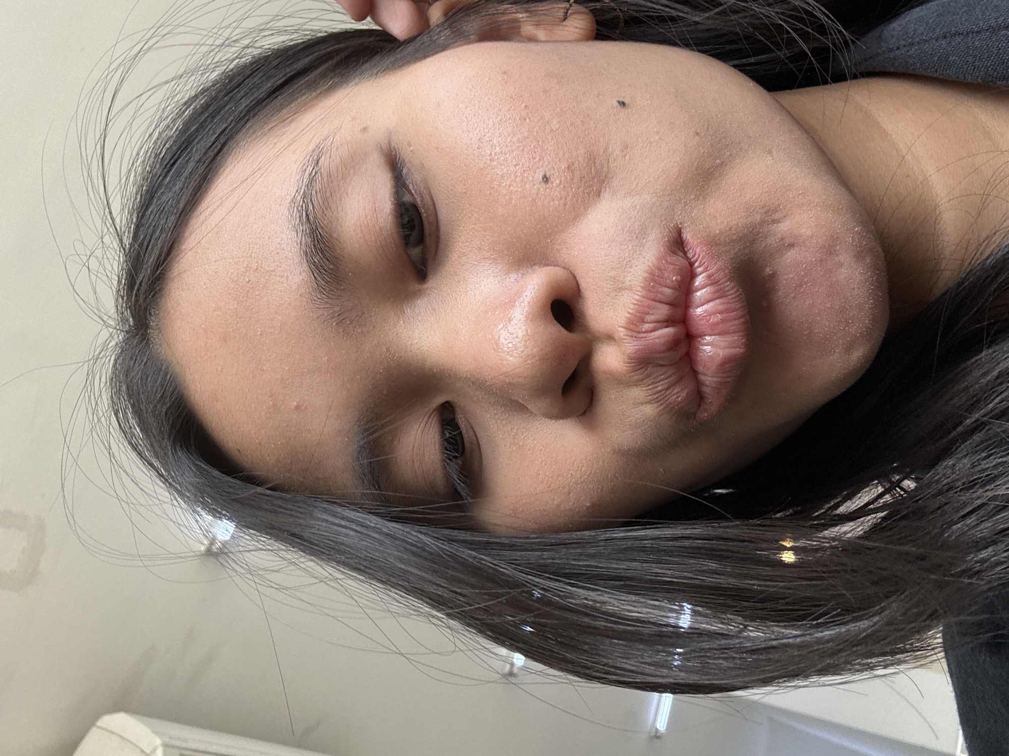 The image is a close-up, low-angle selfie of a young woman with dark hair and brown eyes. She is pouting her lips, and her face is slightly tilted. Her skin appears to have some texture and a few blemishes. There are loose strands of hair framing her face. The background is blurred, but appears to be an indoor setting with fluorescent lighting. There is no specific indication of the location within East Jakarta, Indonesia.