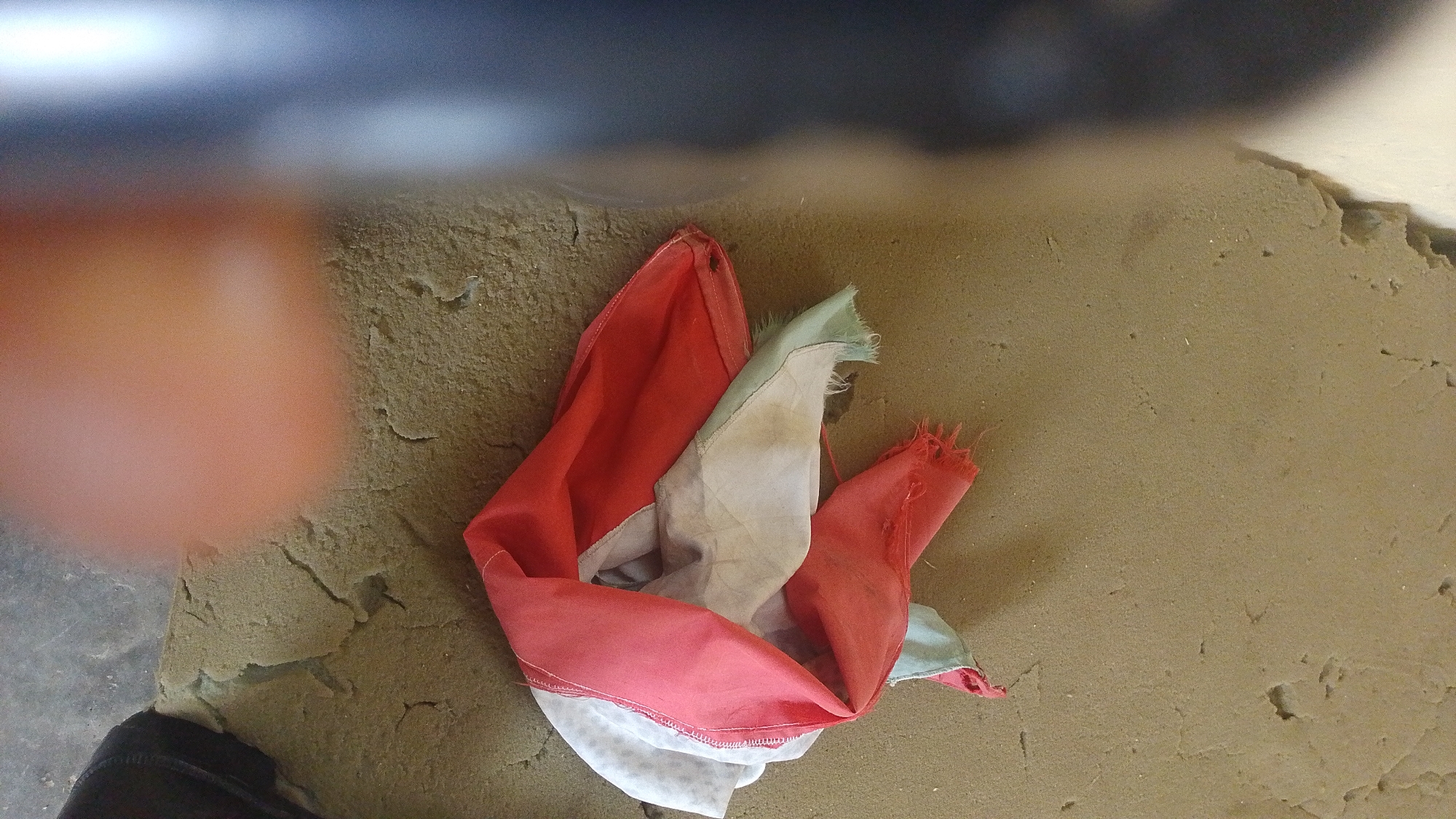 A crumpled red and white fabric lies on a rough, textured, tan surface, possibly a worn floor or an unfinished construction site. The fabric appears to be made of thin material with frayed edges in some areas. A blurred, reddish-orange circular shape, possibly a person's hand or finger, is visible in the upper left quadrant of the image, partially obscuring the background. The upper portion of the image features a dark, out-of-focus area, possibly indicating a ceiling or an overhang. The scene is devoid of any identifying landmarks that would pinpoint a specific location within the city of Balewa, Nigeria. The overall impression is one of disarray and neglect.
