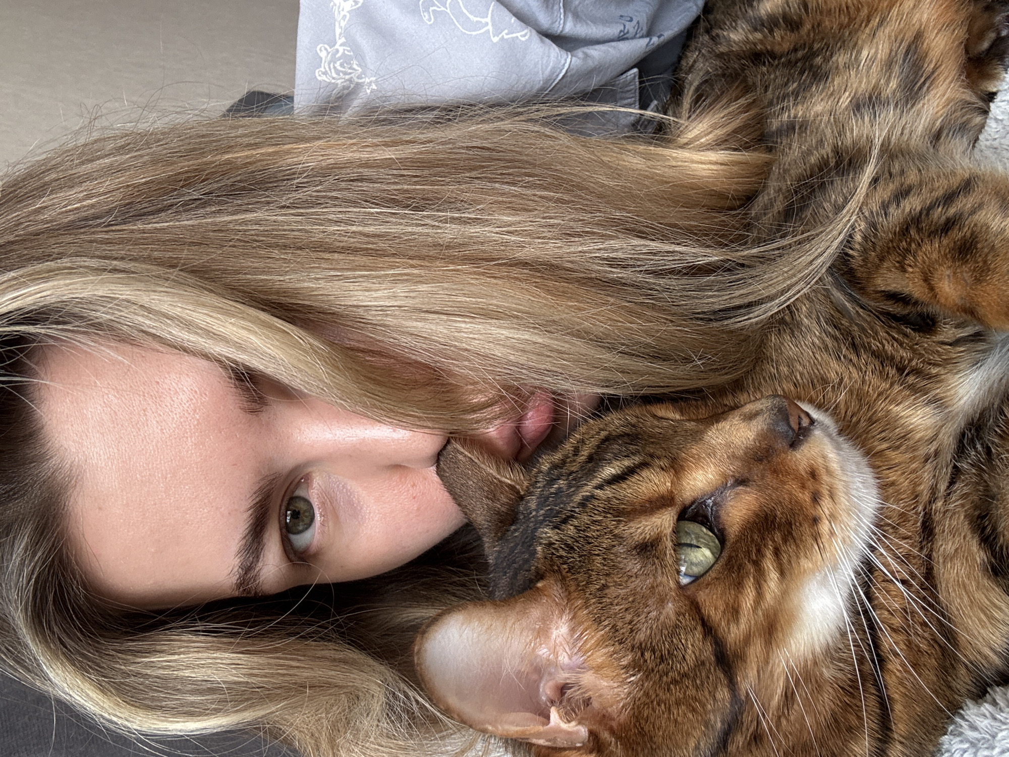 In a close-up, indoor shot, a person with blonde hair is embracing a tabby cat. The person's face is partially obscured by their hair and their cheek is pressed against the cat's head, with their nose touching the cat's ear. The cat, which has green eyes and brown and black stripes, looks towards the left of the frame. The person is wearing a light-colored shirt with a subtle pattern. The background is indistinct, suggesting an indoor setting.