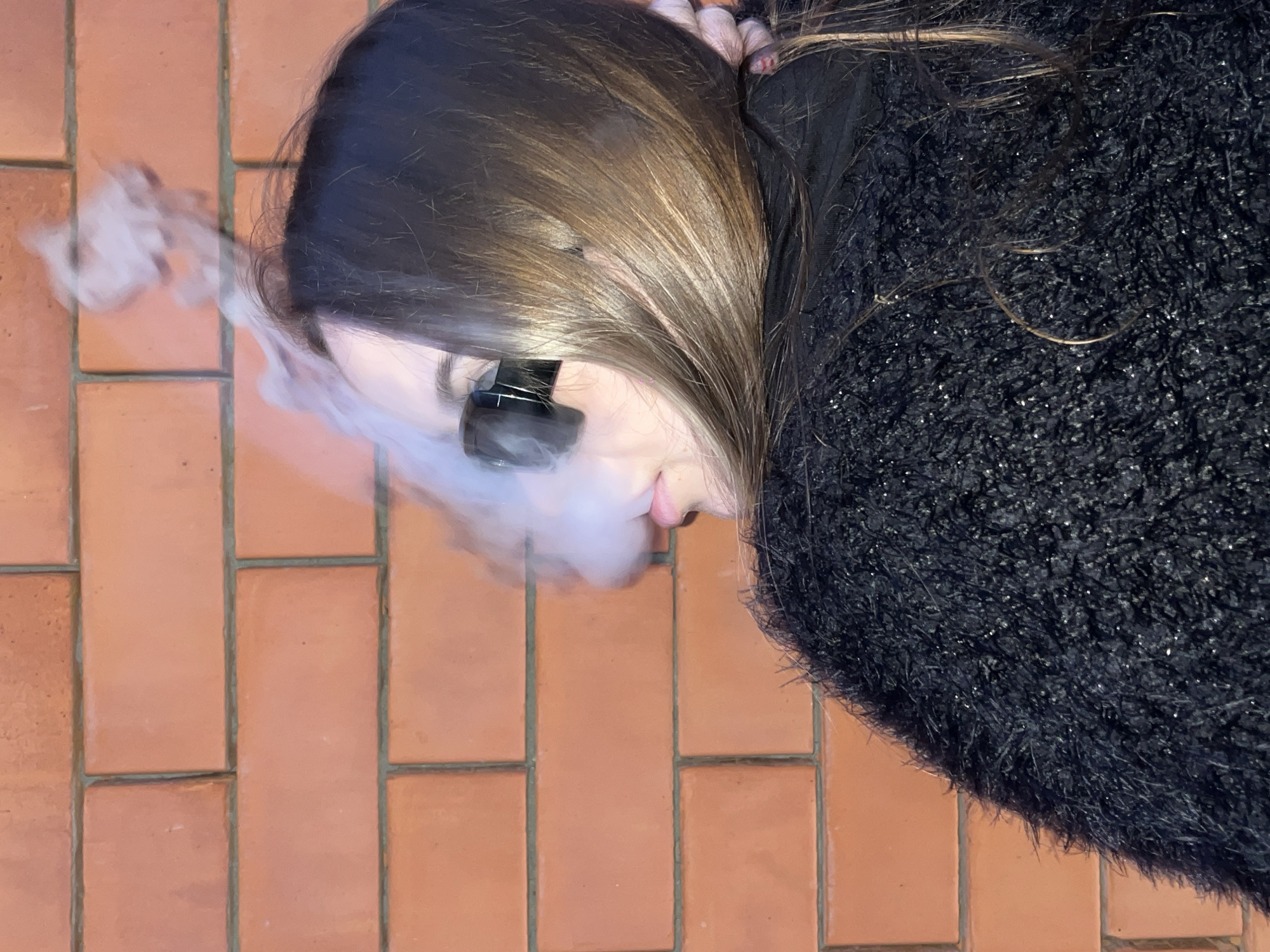 A young woman wearing black fuzzy clothing and dark sunglasses is exhaling a cloud of white vapor in front of a red brick wall. The background is a wall made of orange-red bricks laid in a horizontal pattern. The woman's hair is dark brown and falls over her shoulder. Her lips are slightly parted. The vapor billows upwards and to the left of her head, obscuring part of the brick wall. The image appears to be taken outdoors, likely in a city setting, such as Novopokrovskaya, Russia.