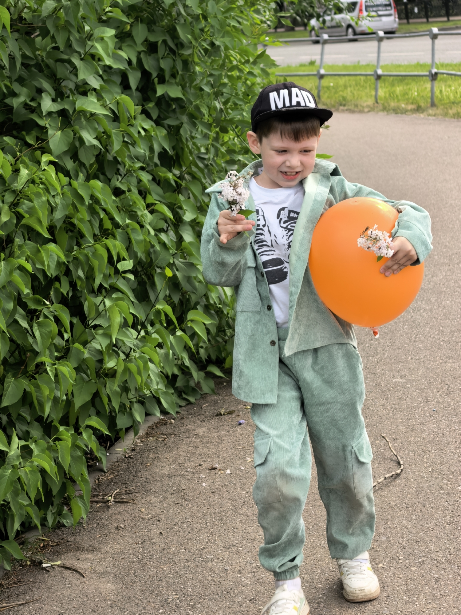 A young boy stands on a paved path next to a lush green bush. He is wearing a black "MAD" baseball cap, a light green corduroy suit, a white t-shirt, and white sneakers. In his right hand, he holds a small bunch of white flowers, and in his left arm, he cradles a large orange balloon decorated with similar white flowers. The boy is looking down with a smile on his face. In the background, a silver van and a metal fence can be seen. The location is Sterlitamak, Russia. A thin stick lies on the pavement near the boy's feet.