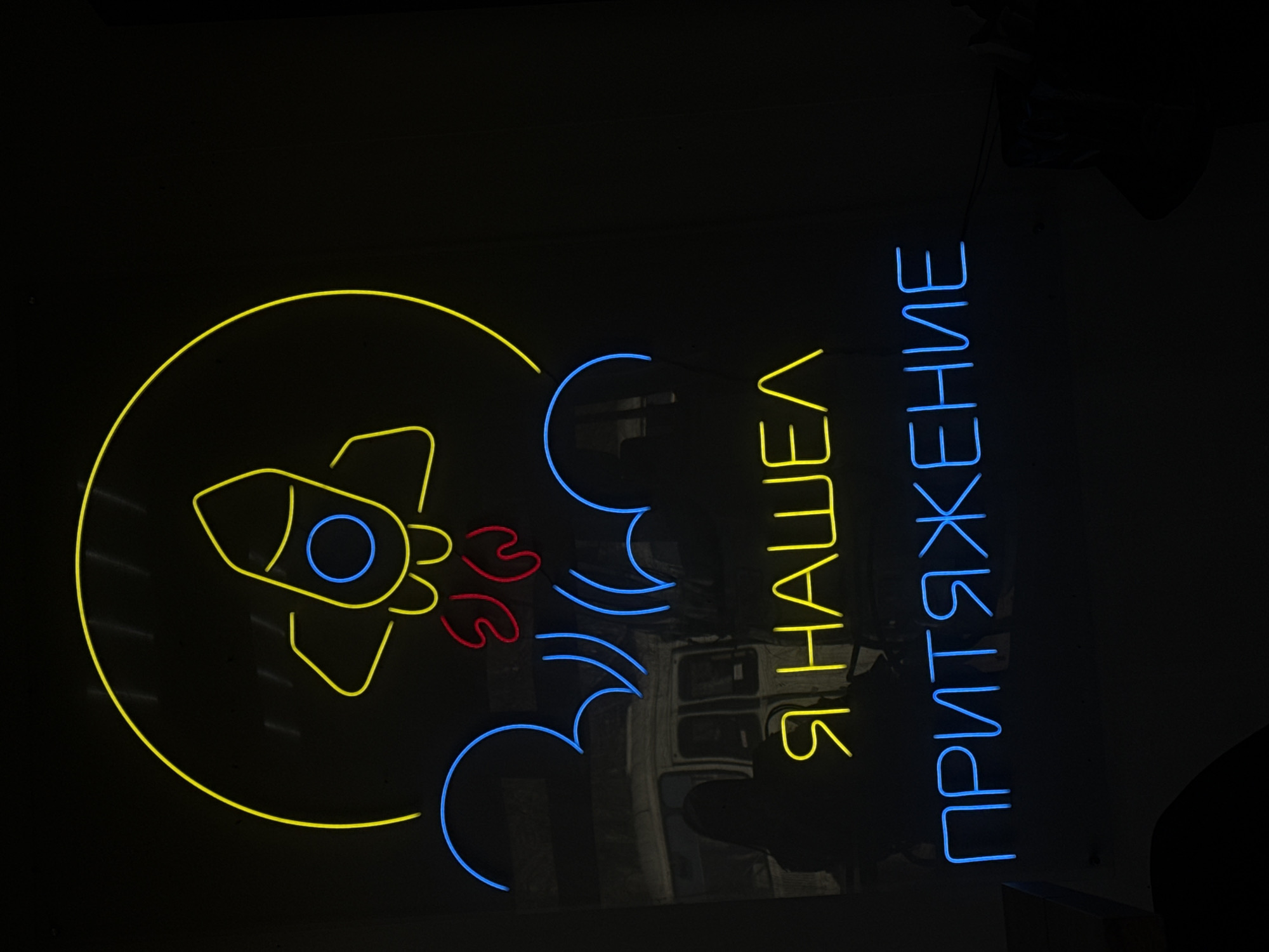 The image shows a neon sign in Votkinsk, Russia. The sign features a yellow rocket inside a yellow circle, blue smoke trails, and red flames. To the right of the rocket is a vertical inscription in Cyrillic script, "Я НАШЕЛ ПРИТЯЖЕНИЕ", lit in blue neon. The background is dark.