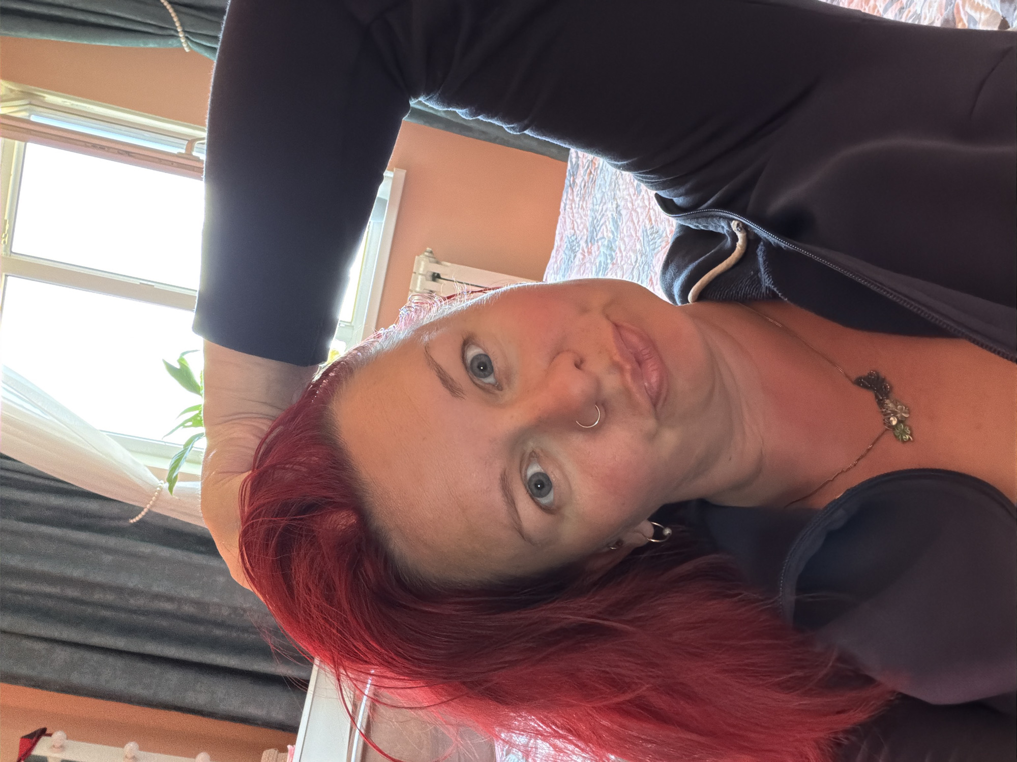 A selfie angled upwards shows a woman with red hair, blue eyes, and a nose ring. She is wearing a dark zippered garment and a necklace. In the background, there's a window with sheer curtains, a plant, and a glimpse of a radiator. A patterned bedspread is also visible. The location in Chaikovsky, Russia is not discernible from the image.