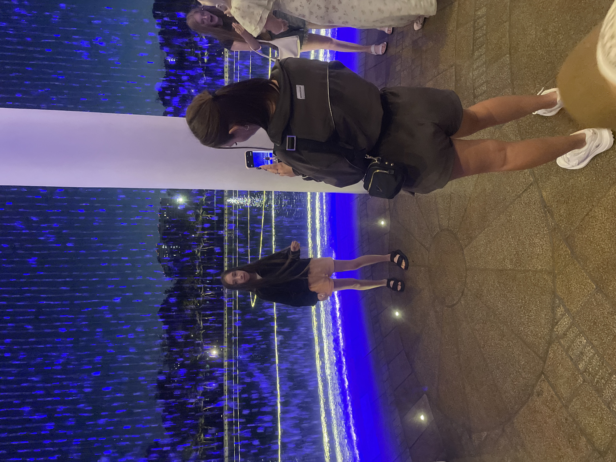 In Krasnodar, Russia, at night, a young girl stands near a illuminated water feature, posing for a photograph. She is wearing a black zip-up jacket, orange shorts, and black sandals. Behind her, a woman is taking her picture with a smartphone. Another woman, with blonde hair, is visible in the background, and a third person, in a white dress, is partially visible on the right. The environment is a paved area with a large, white, cylindrical structure to the right of the girl. The background features a cascading water display illuminated with vibrant blue lights, creating a mesmerizing effect. Trees and distant buildings are faintly visible beyond the water feature. The lighting is dramatic, with strong blue hues dominating the scene.