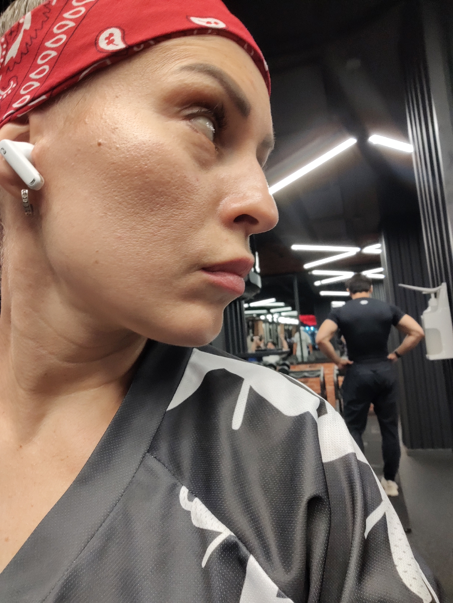 A close-up, head-and-shoulders shot shows a woman from the nose up, wearing a red bandana and a grey shirt with a black and white pattern. She has an earbud in her left ear and is looking off to the right. In the background, a man in a black t-shirt and pants is seen from the back, standing in what appears to be a gym. The gym environment is visible with reflective surfaces and linear lighting. The image does not provide any specific location within Almaty, Kazakhstan.