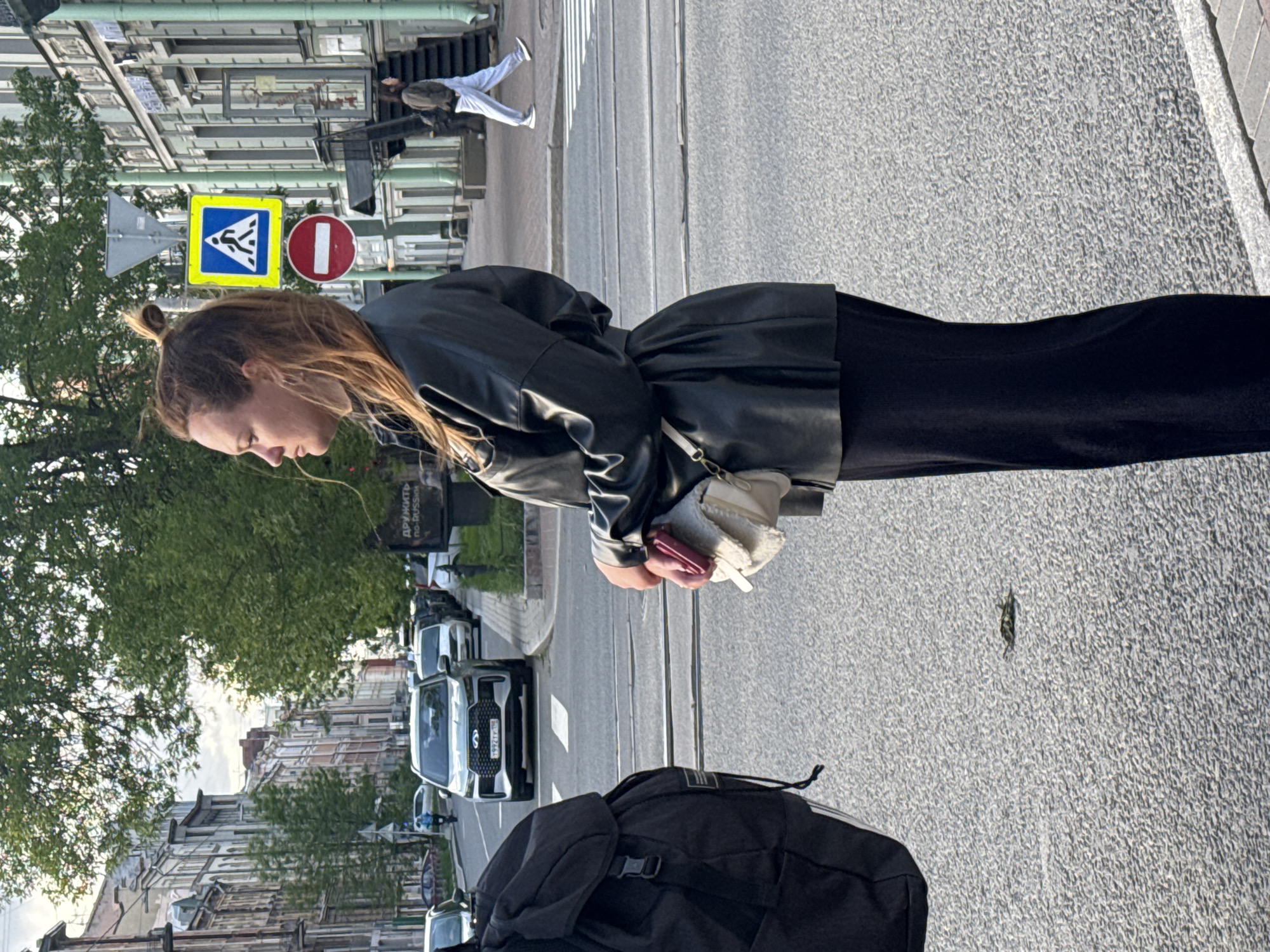 A woman in a black leather jacket stands on a city street with a black backpack in the foreground. She holds a pink object in her hands. The street has light-colored pavement. Traffic flows on the adjacent road, with several cars visible. Buildings line the street, with trees and signage visible along the sidewalk. A pedestrian crosswalk sign and a "do not enter" sign are visible in the background. The street scene suggests the location is in Saint Petersburg, Russia, based on architectural style and signage in Cyrillic script.