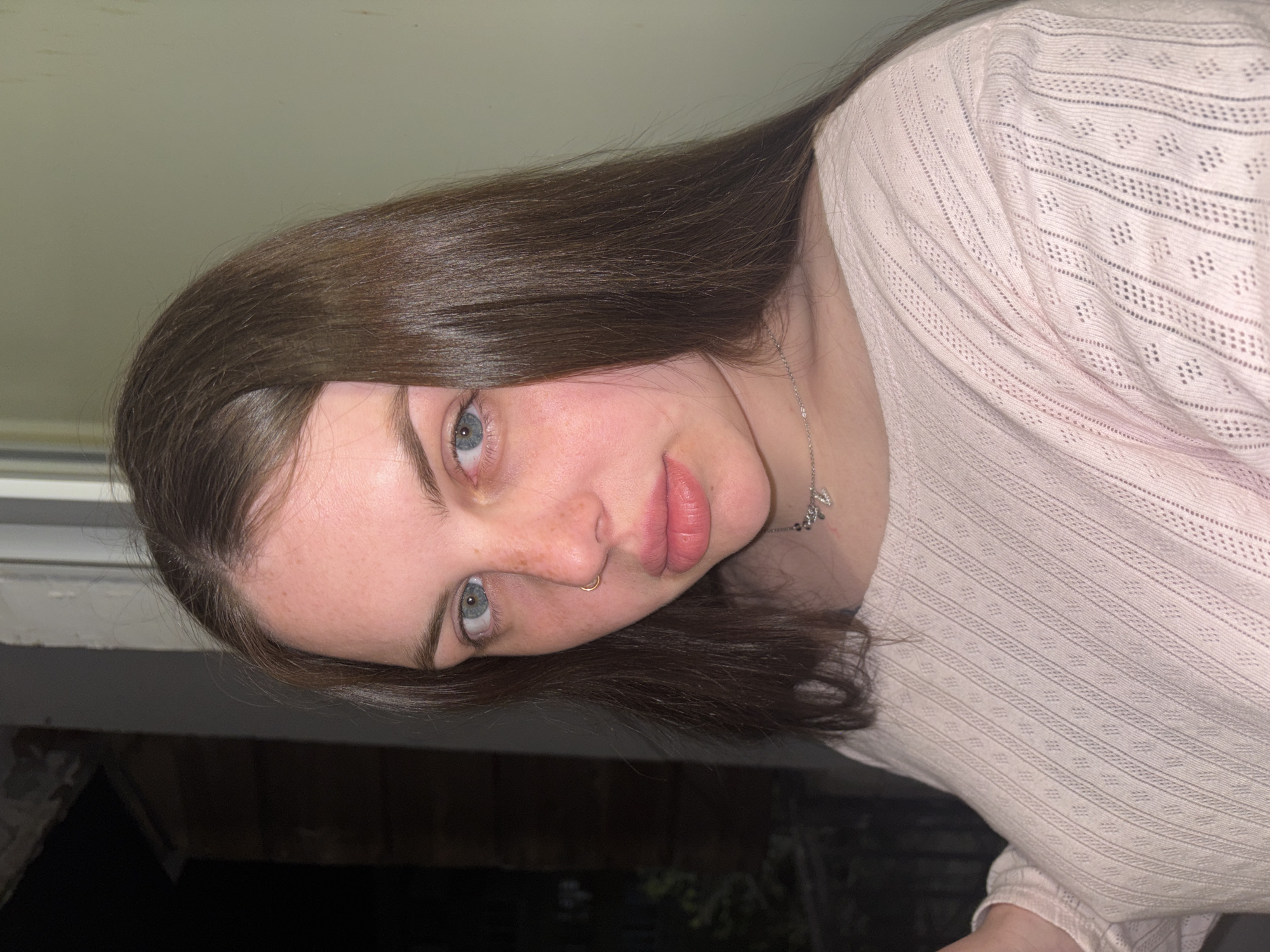 A young woman is depicted in a close-up, waist-up portrait, looking directly at the camera with her head tilted to the right. She has long, straight, glossy dark brown hair, parted on the side, framing her face. Her eyes are a striking light blue or grey, and her skin is fair, accented by noticeable freckles across her nose and cheeks. Her lips are full and appear a natural pink. A small, golden-colored hoop nose ring is visible on her right nostril.
She is wearing a light pink, long-sleeved top, which appears to be made of a textured, open-knit fabric, featuring patterns of horizontal stripes and small, diamond-shaped holes. A thin silver-colored necklace with small, indistinct charms (too small to clearly identify letters or symbols) is visible around her neck.
The background is largely dark and out of focus, suggesting an indoor setting or a nighttime scene. A lighter, neutral-toned surface, possibly a ceiling or wall, is visible above her head in the upper left of the image, while the area below her appears very dark, possibly an unlit room or an outdoor view at night. The lighting illuminates her face, making her features stand out against the subdued background. Her expression is calm and neutral, with a direct gaze at the viewer. The overall impression is a focused, personal portrait.