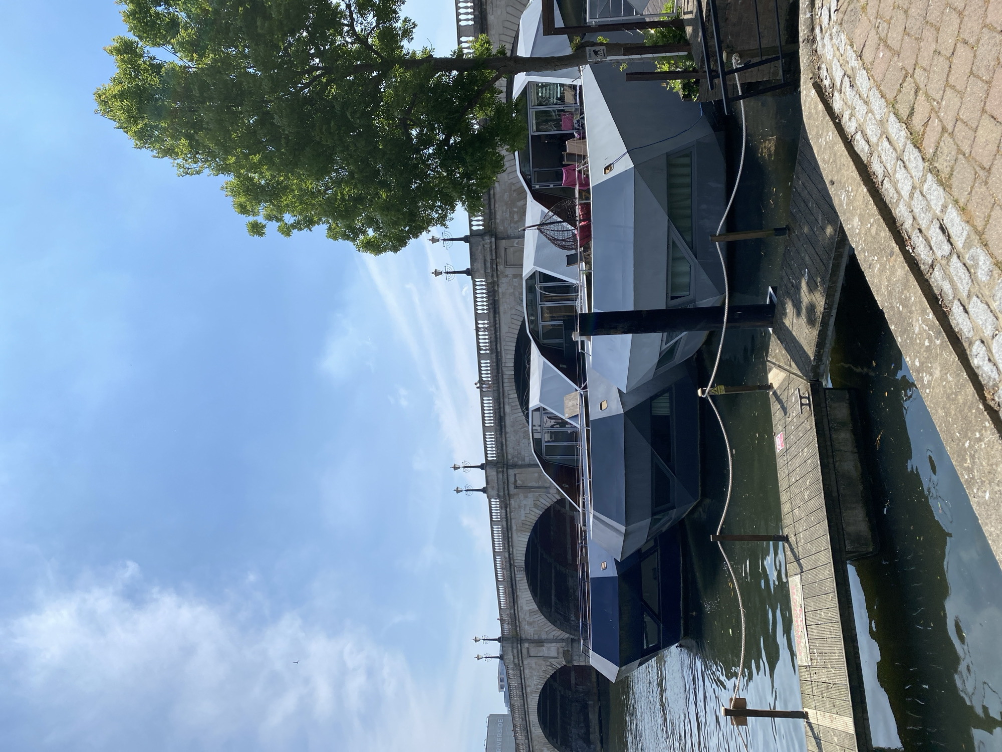 The image depicts a modern house boat moored alongside a quay in London, United Kingdom, with an arched stone bridge forming the backdrop. The boat features geometric paneling in shades of gray and blue, with windows offering a glimpse of interior furnishings. The bridge is constructed of light-colored stone with intricate railings and period lampposts. A mature tree with dense green foliage partially obscures the scene. To the right of the boat, a wooden platform extends over the water, adjacent to a paved pedestrian area. The water is dark and reflective. A clear, blue sky with wispy clouds fills the upper portion of the image.