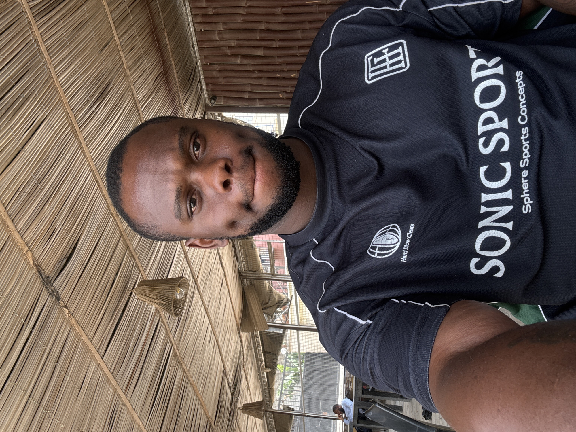 The image is a selfie featuring a dark-skinned man with short dark hair and a beard, looking directly at the camera with a neutral to slightly smiling expression. He is wearing a black short-sleeved athletic shirt with white piping. Visible text on his shirt includes a white logo resembling "IH" or similar letters, and vertically printed "SONIC SPORT" with "Sphere Sports Concepts" beneath it. Below the "IH" logo, another white emblem, possibly a stylized rugby ball, is accompanied by the text "Hard Blow Cleats."
The man is in a semi-open structure, possibly an eatery or lounge, characterized by a striking ceiling made of woven light-colored natural fibers, such as bamboo or palm fronds, creating a thatched or basket-weave pattern. Several hanging light fixtures with shades made of similar woven material are also visible. In the background, hints of an outdoor area or a more open space can be seen through structural openings, including a metal mesh fence. Another person, likely a man in a light-colored shirt, is seated further back in the scene. The natural lighting indicates it is daytime. The setting suggests a casual, possibly rustic, establishment consistent with the location context of Abuja, Nigeria.