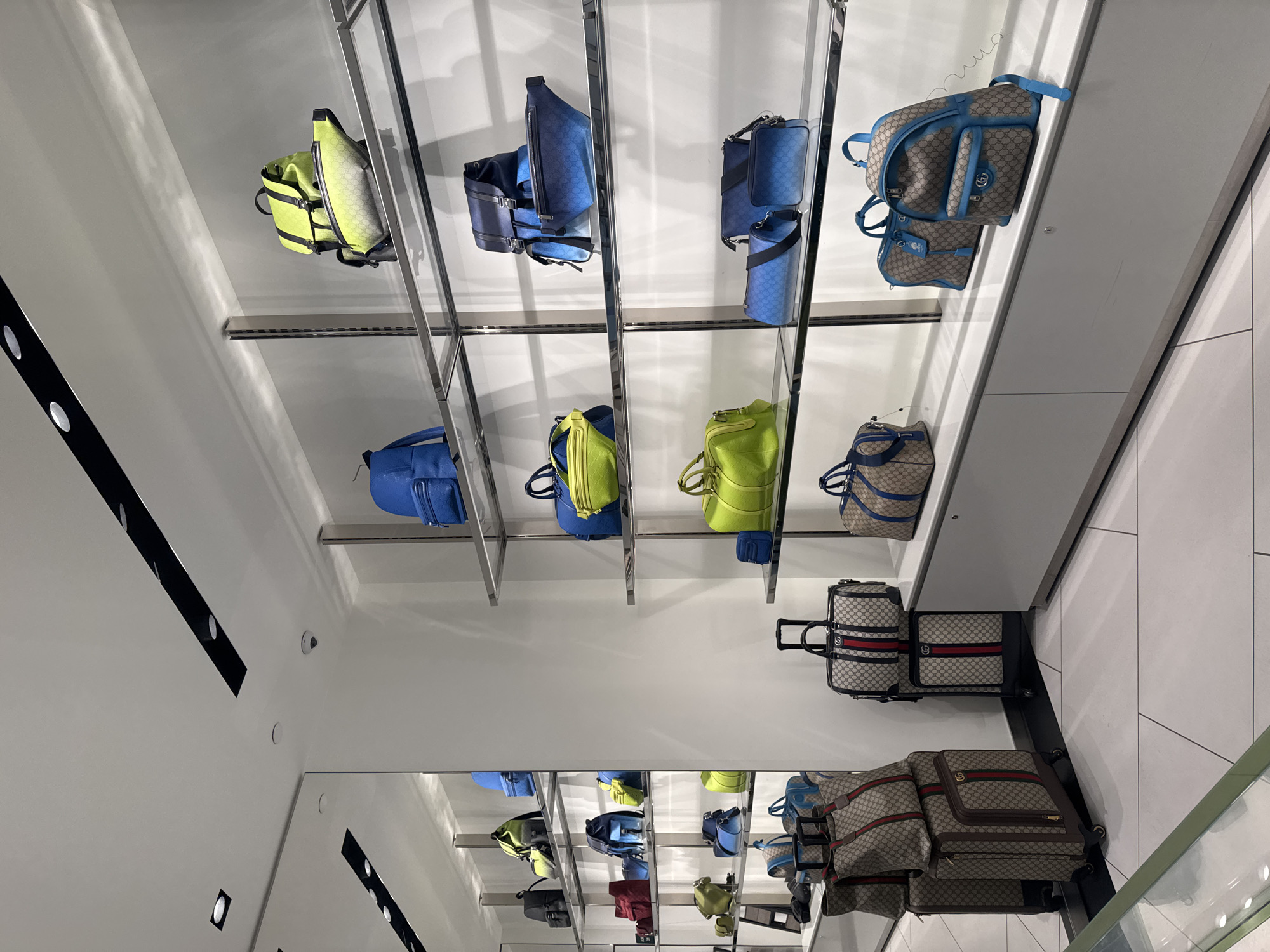 The image displays an indoor retail setting, likely a luxury boutique or department store, showcasing a variety of bags and luggage. The scene is dominated by multiple levels of metal-framed shelves, holding an array of handbags, backpacks, and duffel bags, against bright white walls and a ceiling.

The bags are presented in distinct color schemes. Many smaller bags, including backpacks and cross-body styles, are featured in vibrant bright blue and vivid lime green, often with subtle, tone-on-tone monogram patterns. On the lower shelves and the floor, larger pieces of luggage, such as rolling suitcases and duffel bags, exhibit a classic beige and brown monogram pattern, frequently accented with the iconic red and green striped webbing, characteristic of the Gucci brand.

No people are visible in the image; the scene is static, focused entirely on the product display. The store environment is brightly illuminated by recessed lighting fixtures embedded in the ceiling and along the walls. The floor is covered with light-colored tiles, and a large mirror or reflective surface is visible towards the bottom, showing reflections of the display and overhead lights. The overall impression is one of a clean, modern, and high-end retail space designed to showcase luxury accessories. While no explicit brand name is visible, the distinctive patterns on the bags provide strong brand identification.