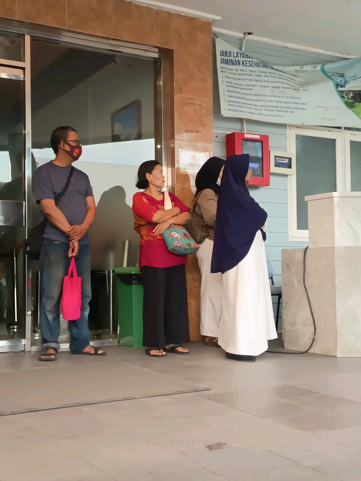 The image depicts a scene outside a building, possibly a health clinic or hospital, in Tangerang, Indonesia. Several people are standing near an entrance.
In the foreground, a man wearing a dark blue t-shirt, jeans, glasses, and a face mask stands on the left. He has a black shoulder bag and is holding a bright pink tote bag in his right hand. Next to him, a woman in a red top with an orange and brown patterned overlay and black pants stands with her arms crossed. She has a patterned tote bag slung over her right shoulder.
Behind them, two women are partially visible. One wears a light brown hijab and a checkered bag. The other is dressed in a dark blue hijab and a long white garment.
A green trash can is situated to the left of the entrance. Above the people, a banner with text in Indonesian is displayed, which appears to be a service pledge. The building's facade features tiled walls and glass doors. The ground is paved with grey tiles.