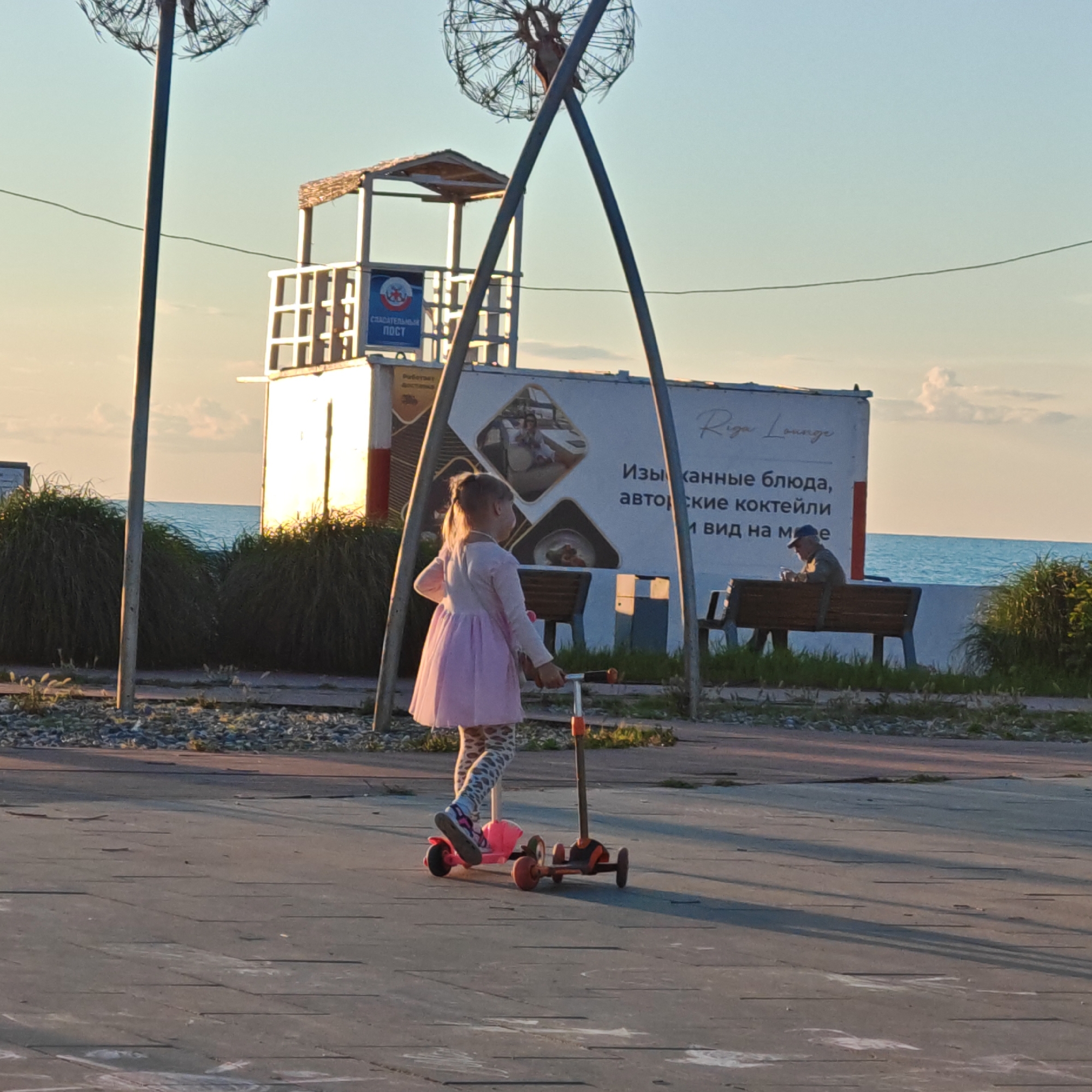 An eye-level shot captures a vibrant coastal scene at what appears to be late afternoon or early evening, characterized by warm, golden light and long shadows.
In the foreground, a young girl with her hair in a ponytail, wearing a pink dress and patterned leggings, stands on a paved surface next to a pink and orange scooter. Her left foot is on the scooter, suggesting she is about to ride or has just paused.
Behind the girl, several tall, dark metal poles rise, supporting large, intricate structures that resemble giant dandelion seed heads. To the right of the girl, the poles form an A-frame. Further back, a white, two-story structure, identified by a blue sign as a "СПАСАТЕЛЬНЫЙ ПОСТ" (Lifeguard Post), stands near the water. Adjacent to this is a large billboard advertising "Riga Lounge" with Russian text that reads: "Изысканные блюда, авторские коктейли и вид на море" (Exquisite dishes, signature cocktails, and sea view).
To the right of the billboard, an older man wearing a cap is seated on a wooden bench, looking down at something held in his hands, possibly a phone. Tall, reedy green grasses line the edge of the promenade, separating it from the sea.
In the background, the calm blue sea stretches towards the horizon under a light blue, almost cloudless sky, contributing to a serene and recreational atmosphere. The overall impression is one of leisure and relaxation by the coast.
