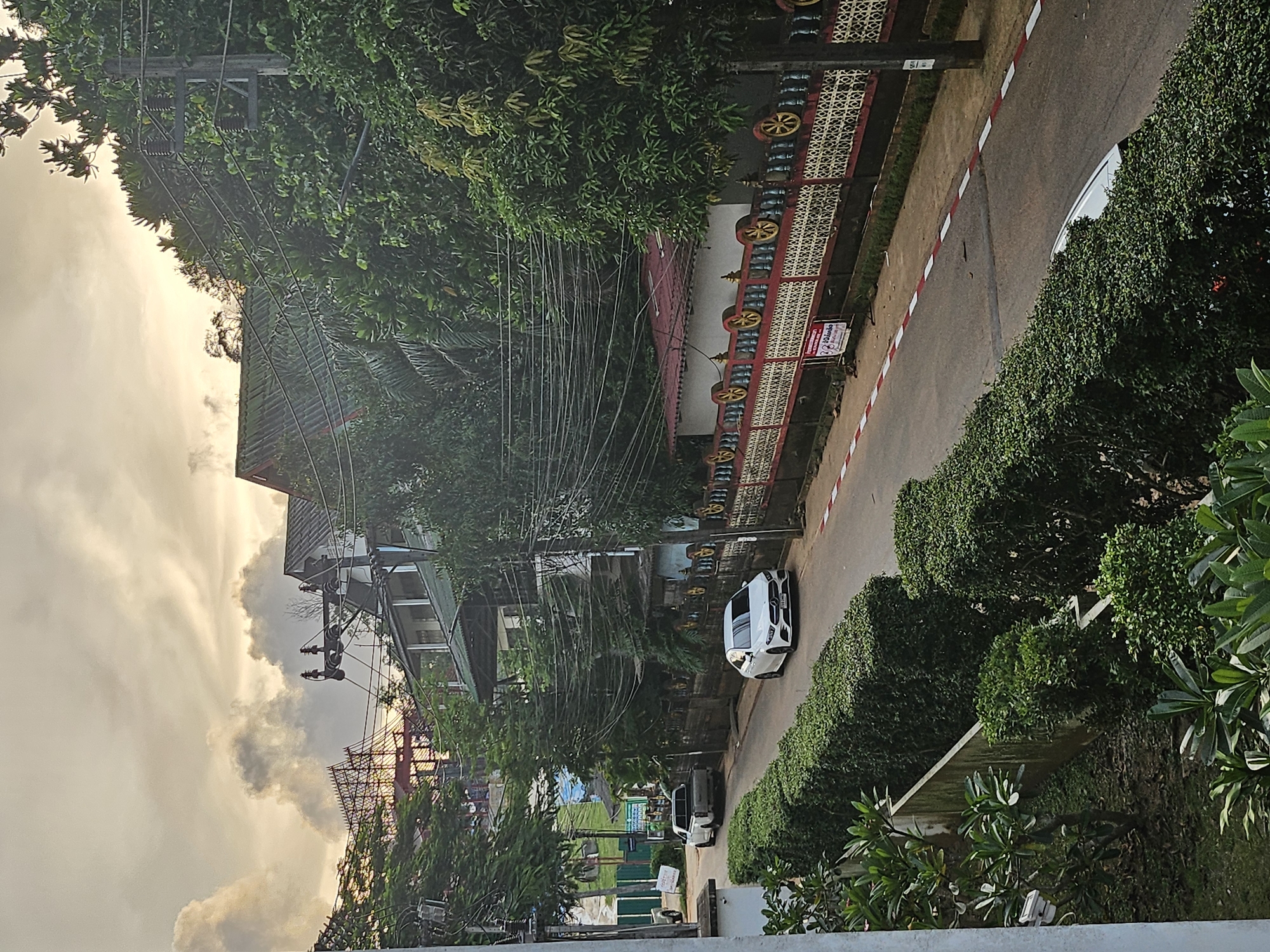 The image depicts a street scene in Kathu, Thailand, under a cloudy sky with golden light filtering through. A paved road runs through the scene, lined with lush green hedges on the left. On the right, a decorative fence with a red and yellow pattern separates the road from buildings and dense foliage. A white Mercedes-Benz sedan is parked on the right side of the road, and a white pickup truck is parked further down on the left. Buildings with green roofs are visible behind the hedges and the fence, with electrical wires crisscrossing the scene. Trees and vegetation are abundant throughout the image.