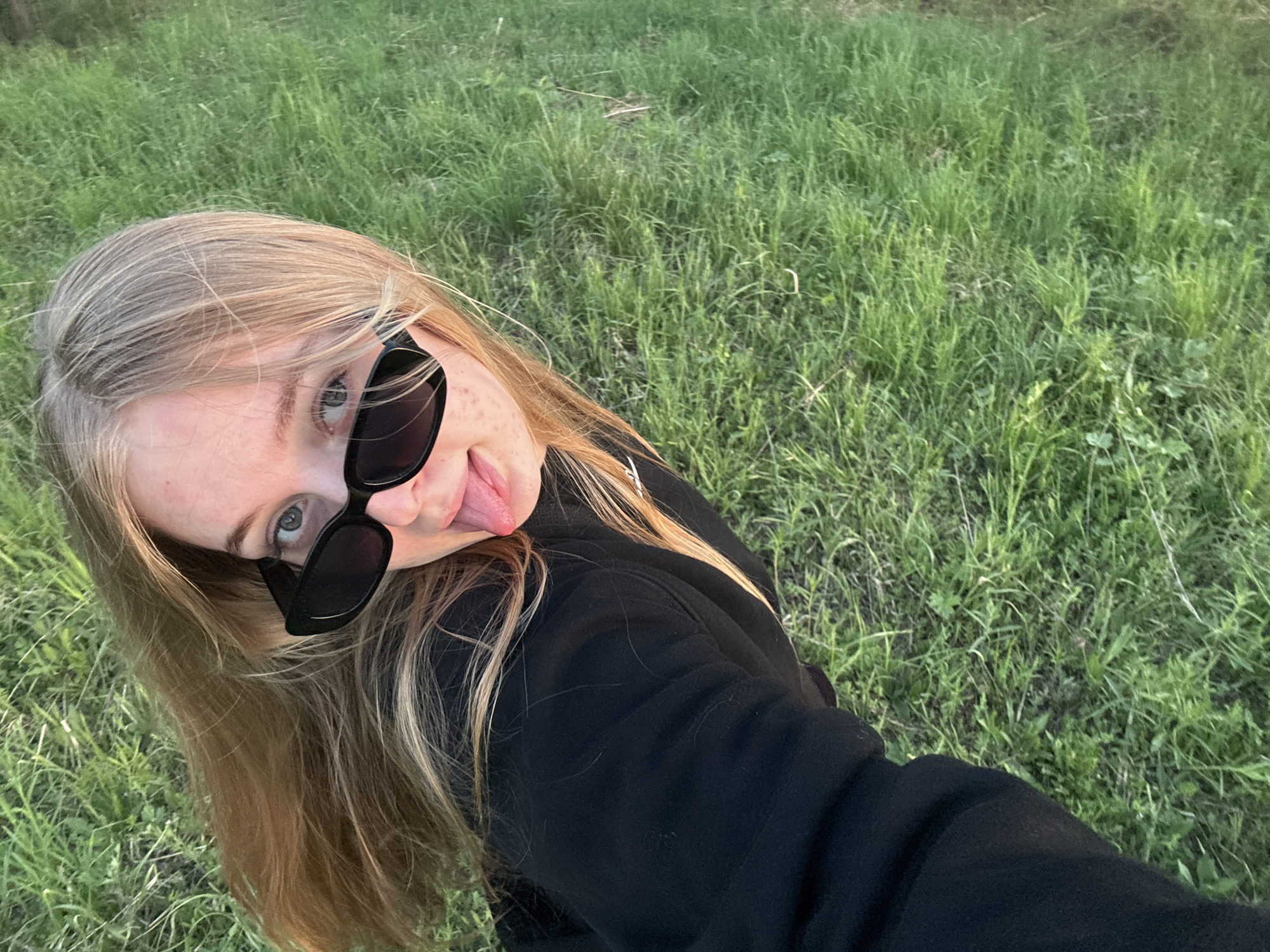 A selfie of a young woman with fair skin and blonde hair, wearing black sunglasses, and a black sweatshirt, sticking her tongue out. The background is a grassy field. The location is unspecified and the description does not specify the city of Zhigalki, Russia.