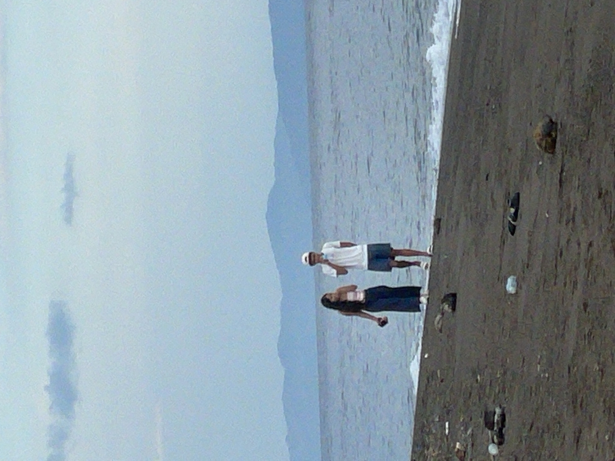The image depicts a beach scene in Dauin, Philippines, with two individuals walking near the water's edge. The ground is dark sand, scattered with debris including plastic bottles and what appear to be flip-flops. The ocean is calm with gentle waves breaking on the shore. In the background, a mountain range is visible under a hazy, overcast sky. One person, a female, is wearing a strapless top and a long, dark skirt. The other person, a male, is wearing a white t-shirt, dark shorts, and a white cap. He is holding a light-colored object, possibly a phone, near his face. The overall lighting suggests it is either early morning or late afternoon.