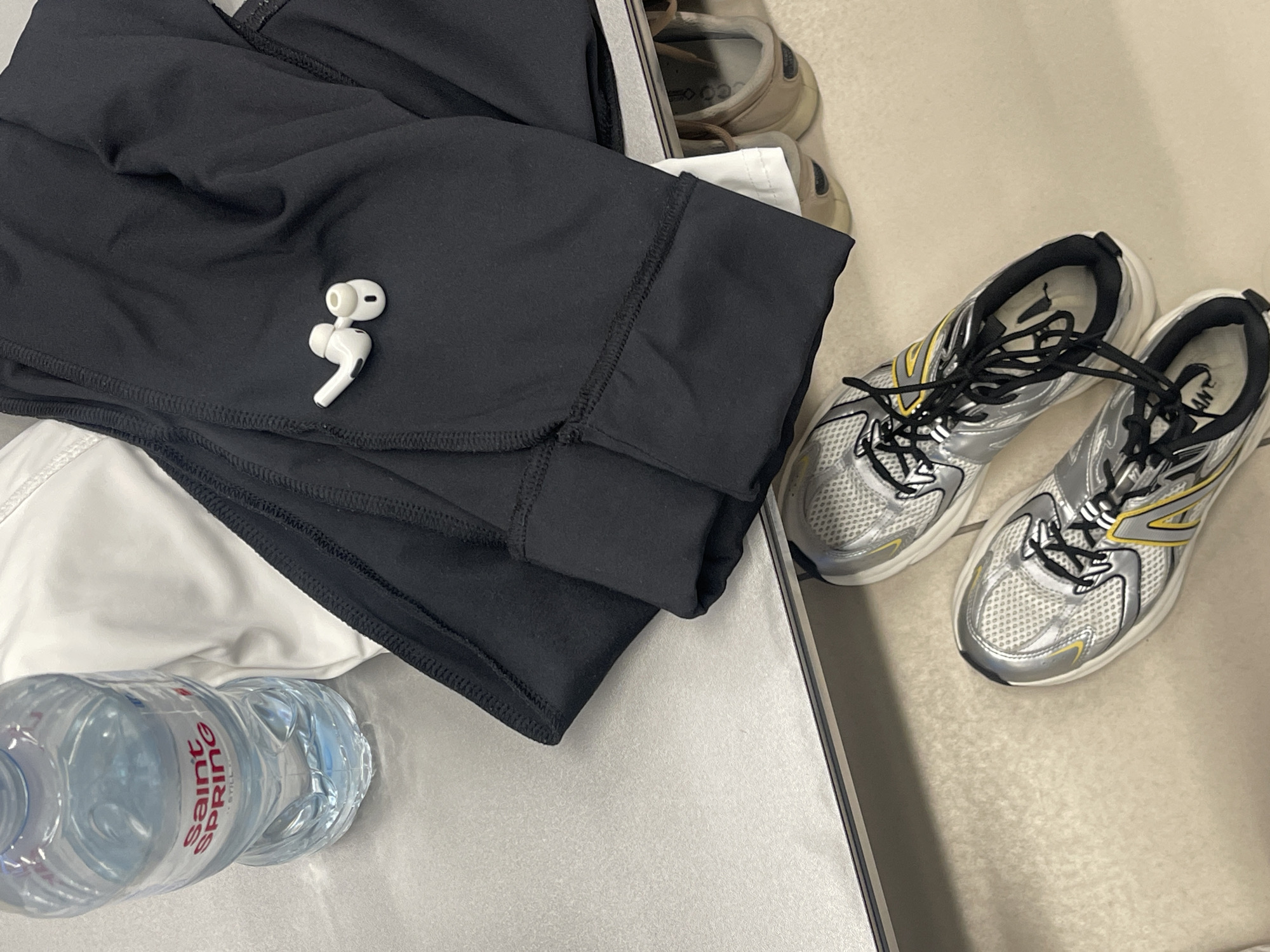 The image depicts a close-up, overhead view of gym or athletic gear laid out on a textured, light gray surface, likely a bench or table. A partially filled plastic water bottle with a red and white "SAINT SPRING" label is positioned on the left. Next to it, folded black athletic pants are visible, with a pair of white wireless earbuds resting on top of the pants. Beneath the table or bench, a pair of silver and yellow athletic shoes are placed, with another pair of shoes, appearing to be slip-on, brown clogs, partially visible further back. The overall scene suggests preparation for or aftermath of physical activity, and the environment appears to be indoors, possibly a locker room or gym. There is no indication of a specific location in the city of Minsk, Belarus.