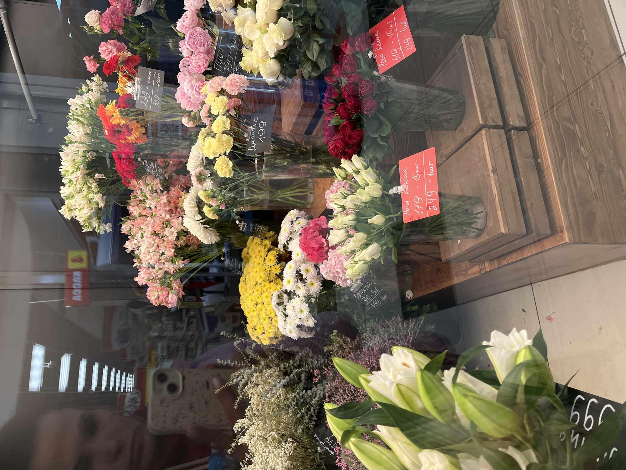 The image depicts a flower shop window in Petropavlovsk-Kamchatsky, Russia. Various bouquets of flowers are displayed in clear vases on wooden shelves. Roses, lilies, carnations, hydrangeas, peonies, and other mixed floral arrangements are visible. Price tags in Russian indicate the cost of individual stems or small bouquets. Reflections in the window show the interior of a store, shelves stocked with merchandise, and a person holding a phone.